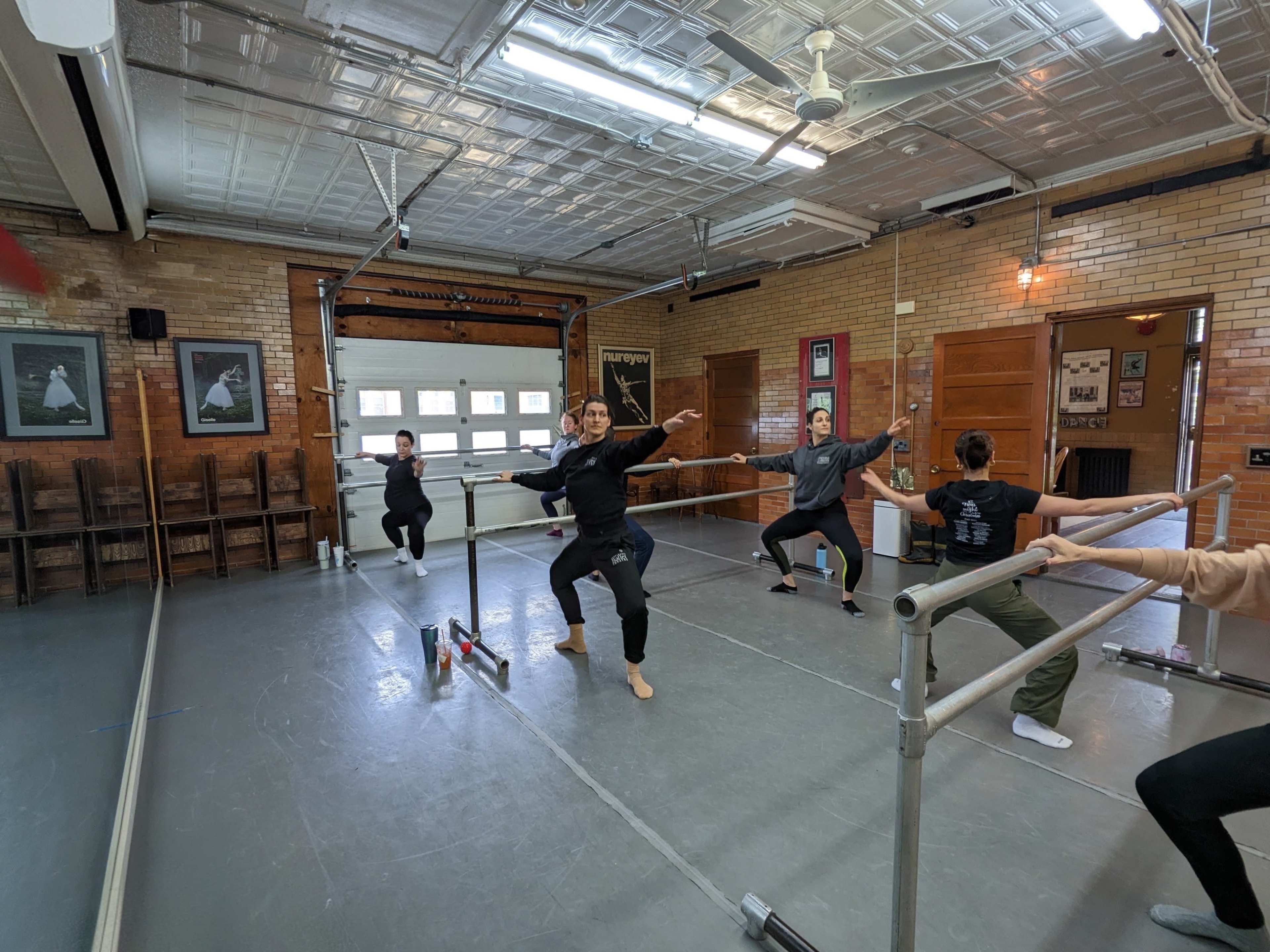 A group of dancers practices ballet in a studio, using a dance bar for support.
