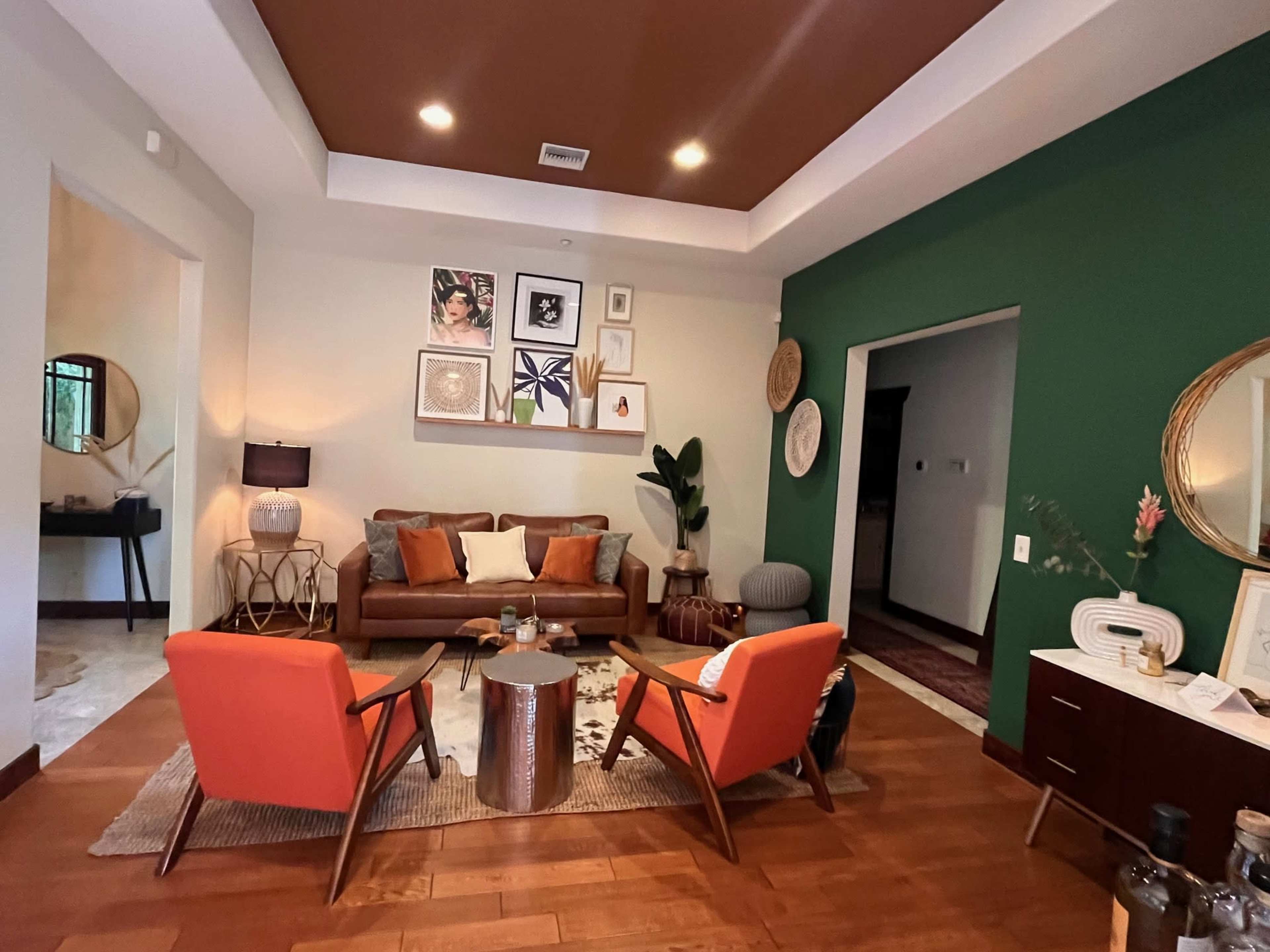 A living room features a brown sofa, two orange chairs, and a dark green accent wall with framed artwork.