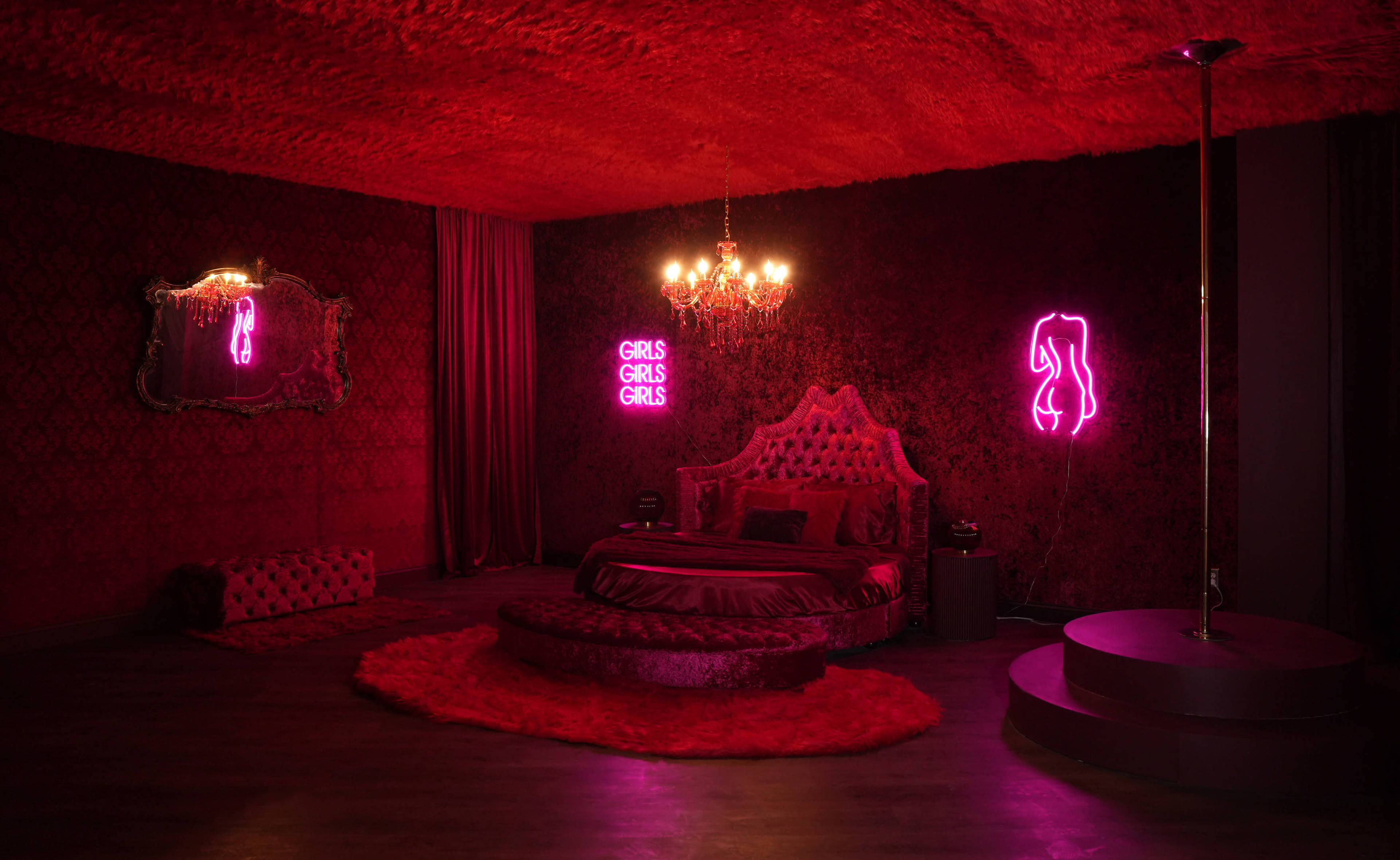 A richly decorated bedroom featuring red velvet walls, a large bed with ornate headboard, and neon signs that read "GIRLS GIRLS GIRLS."