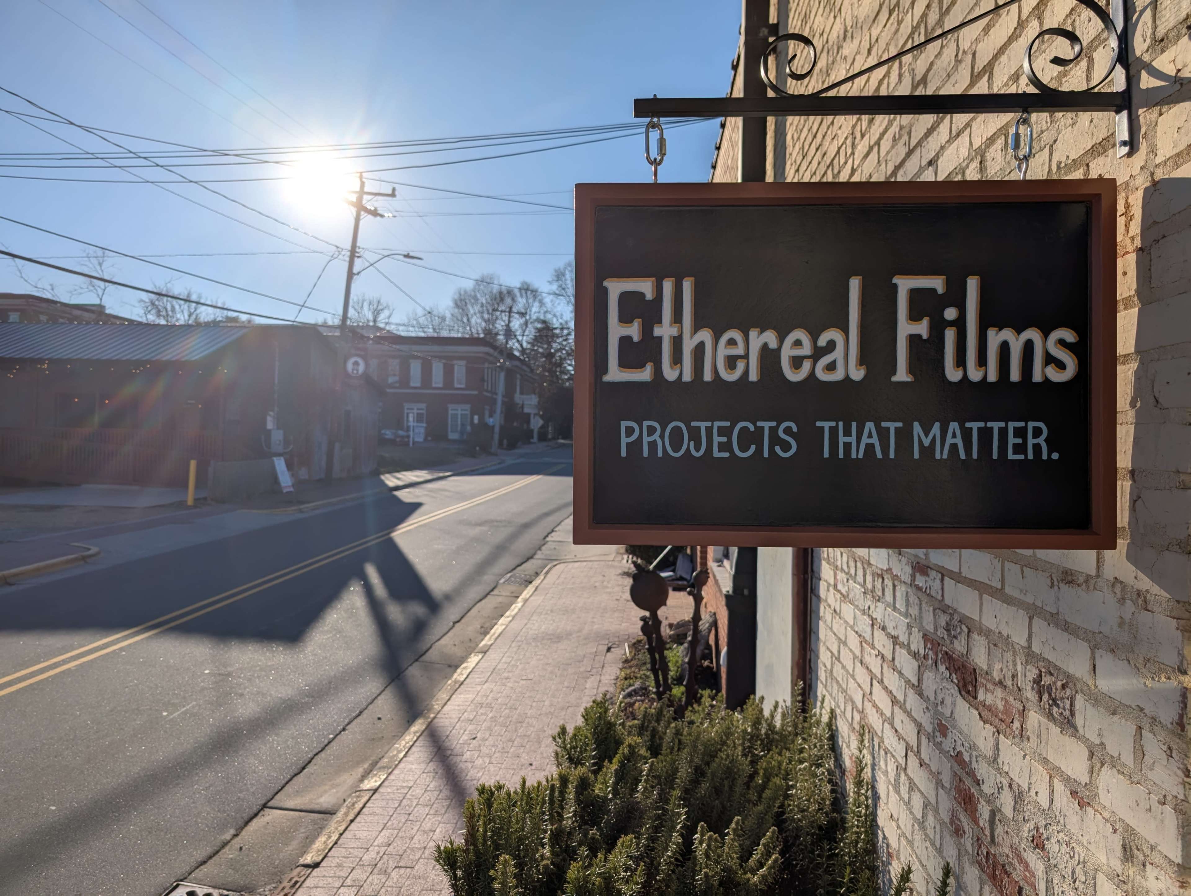 A wooden sign reading "Ethereal Films" hangs outside a building, with sunlight shining in the background and a street visible in the distance.