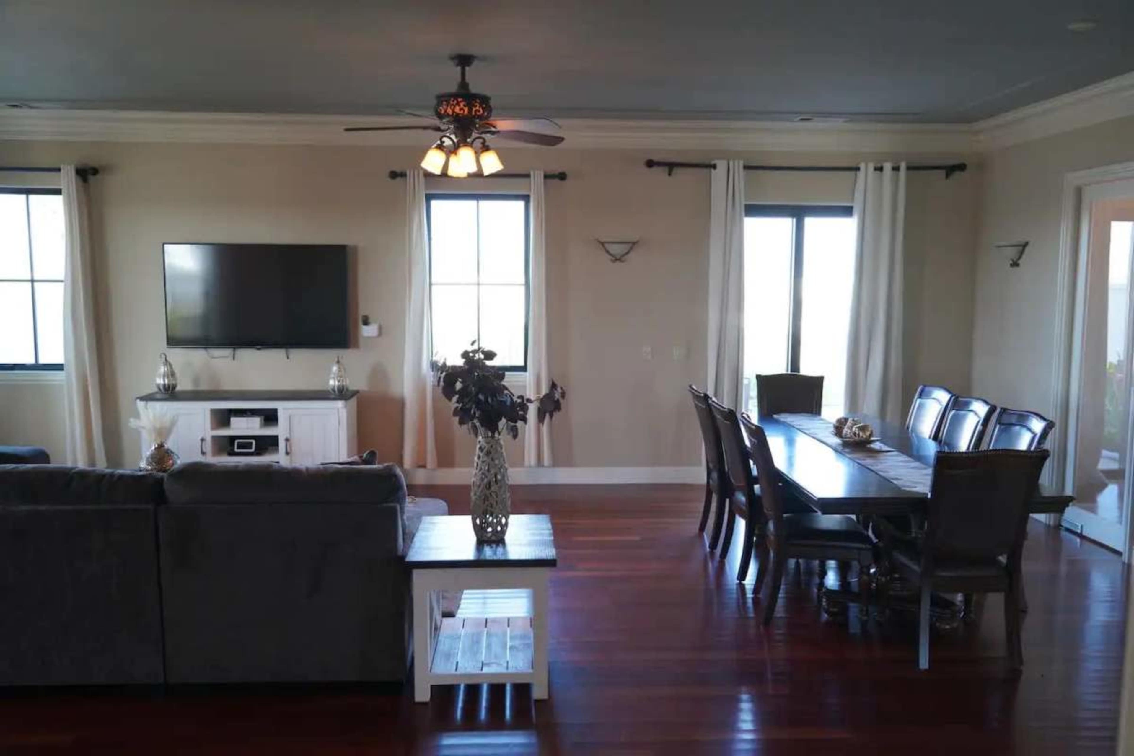 The image shows a spacious living and dining area with hardwood floors, a large table, a television, and light-colored curtains at the windows.