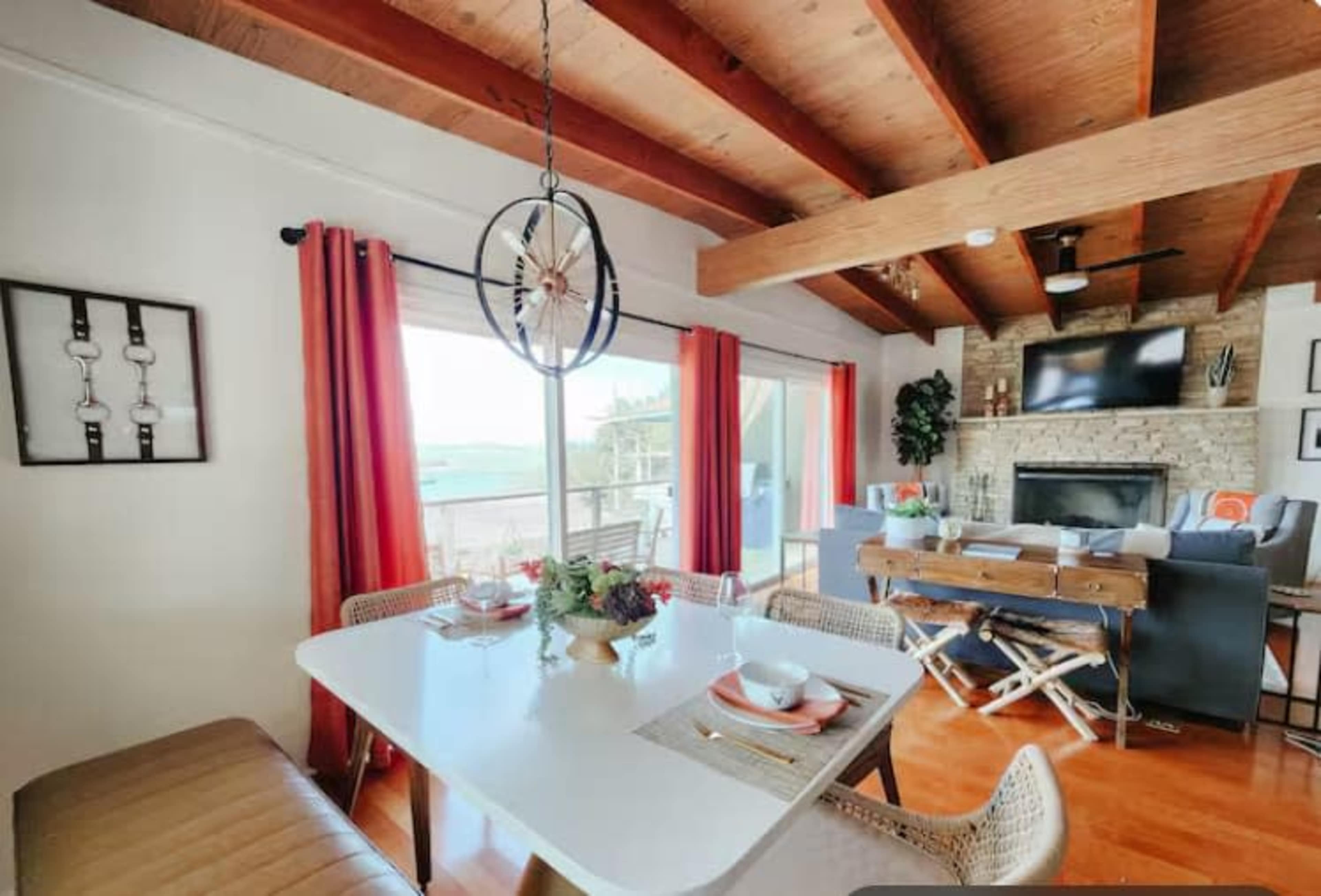 A dining area features a white table set with dishware, surrounded by a beige bench and chairs, with a stone fireplace and large windows visible in the background.