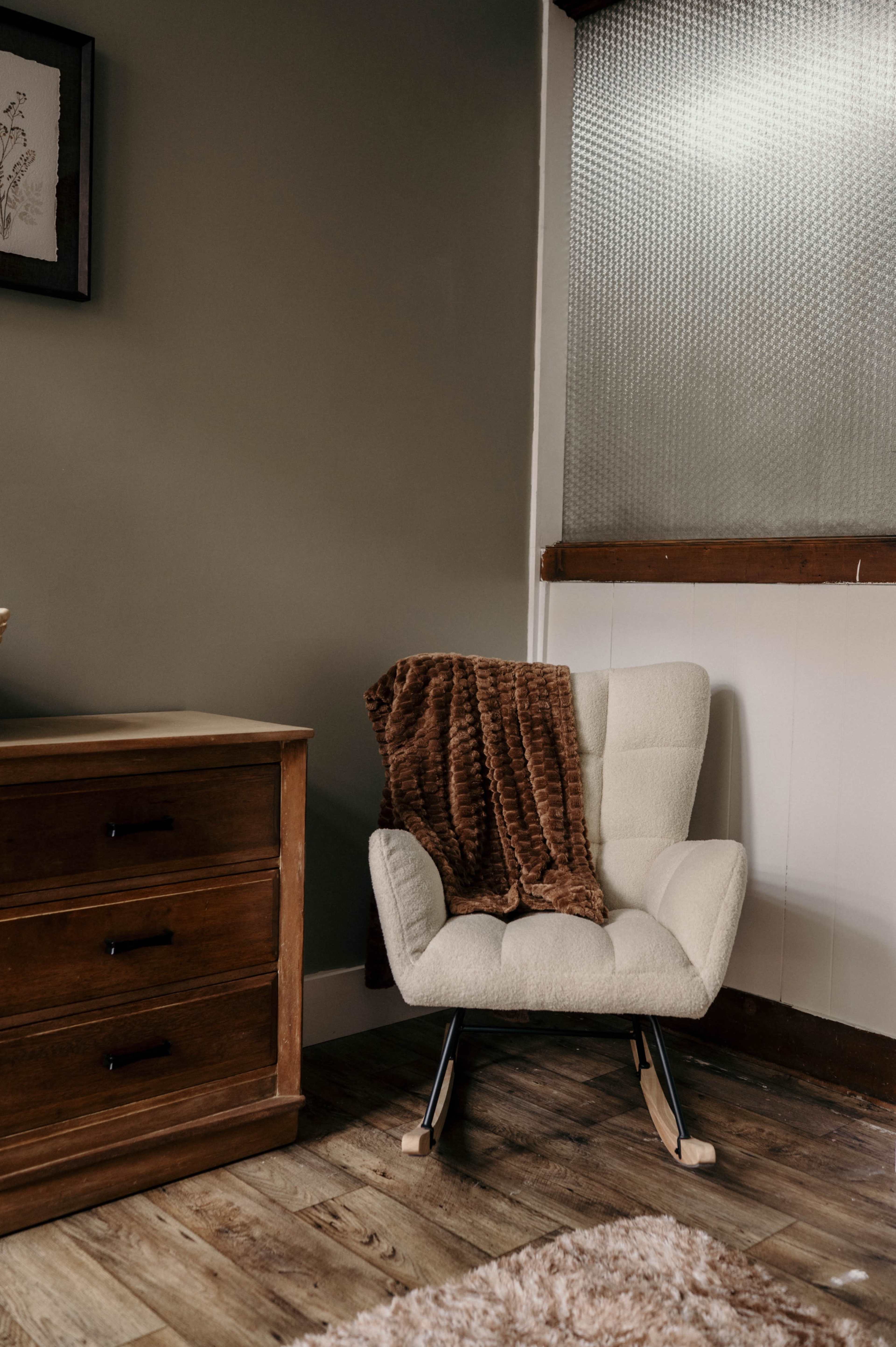 The image shows a cozy corner of a room featuring a white upholstered chair with a brown throw blanket draped over it, adjacent to a wooden dresser.