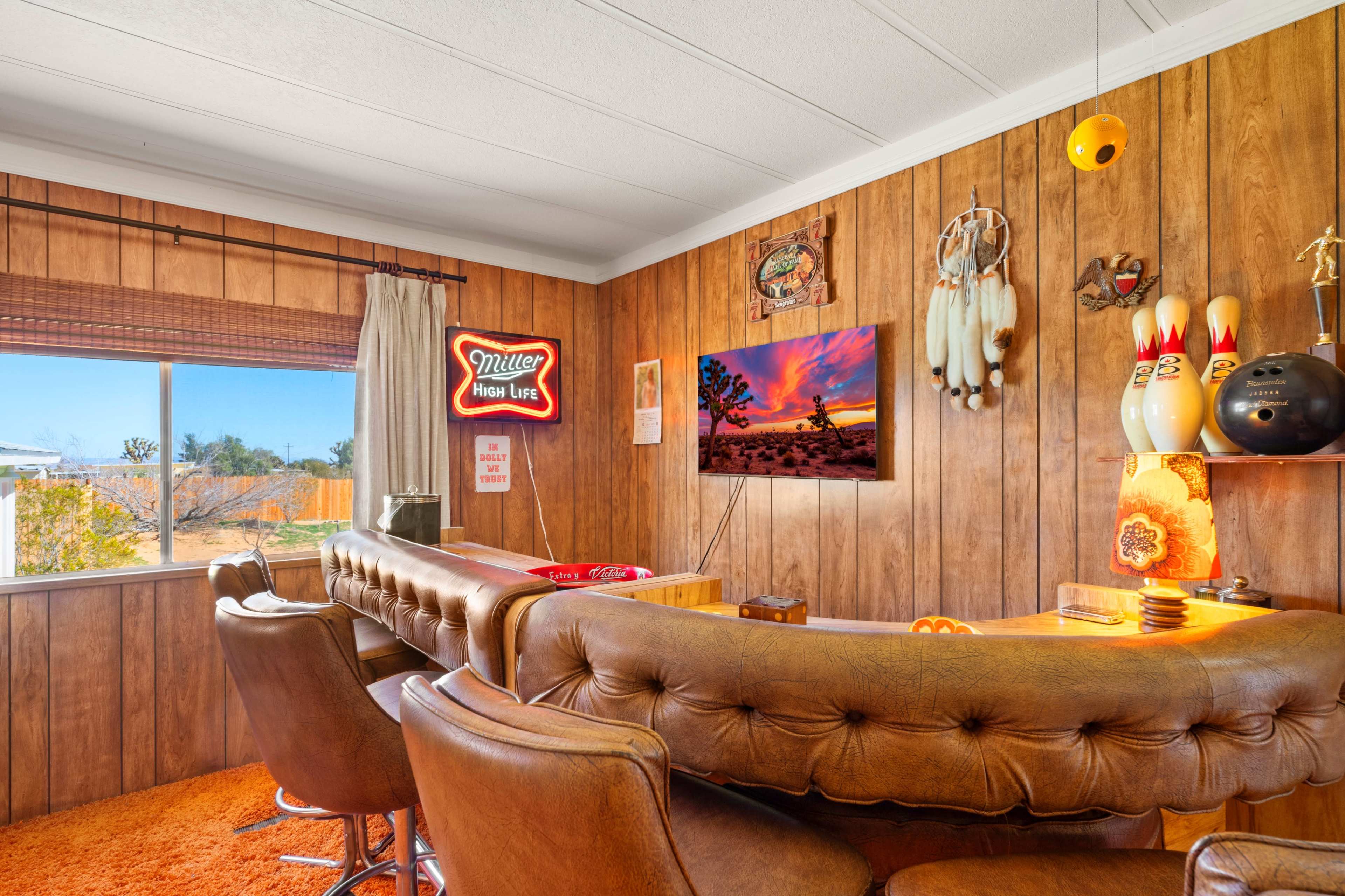 The image shows a cozy room with wooden paneling, a bar area featuring leather chairs, a television displaying a colorful landscape, and decorative items including a bowling pin display and a neon sign.
