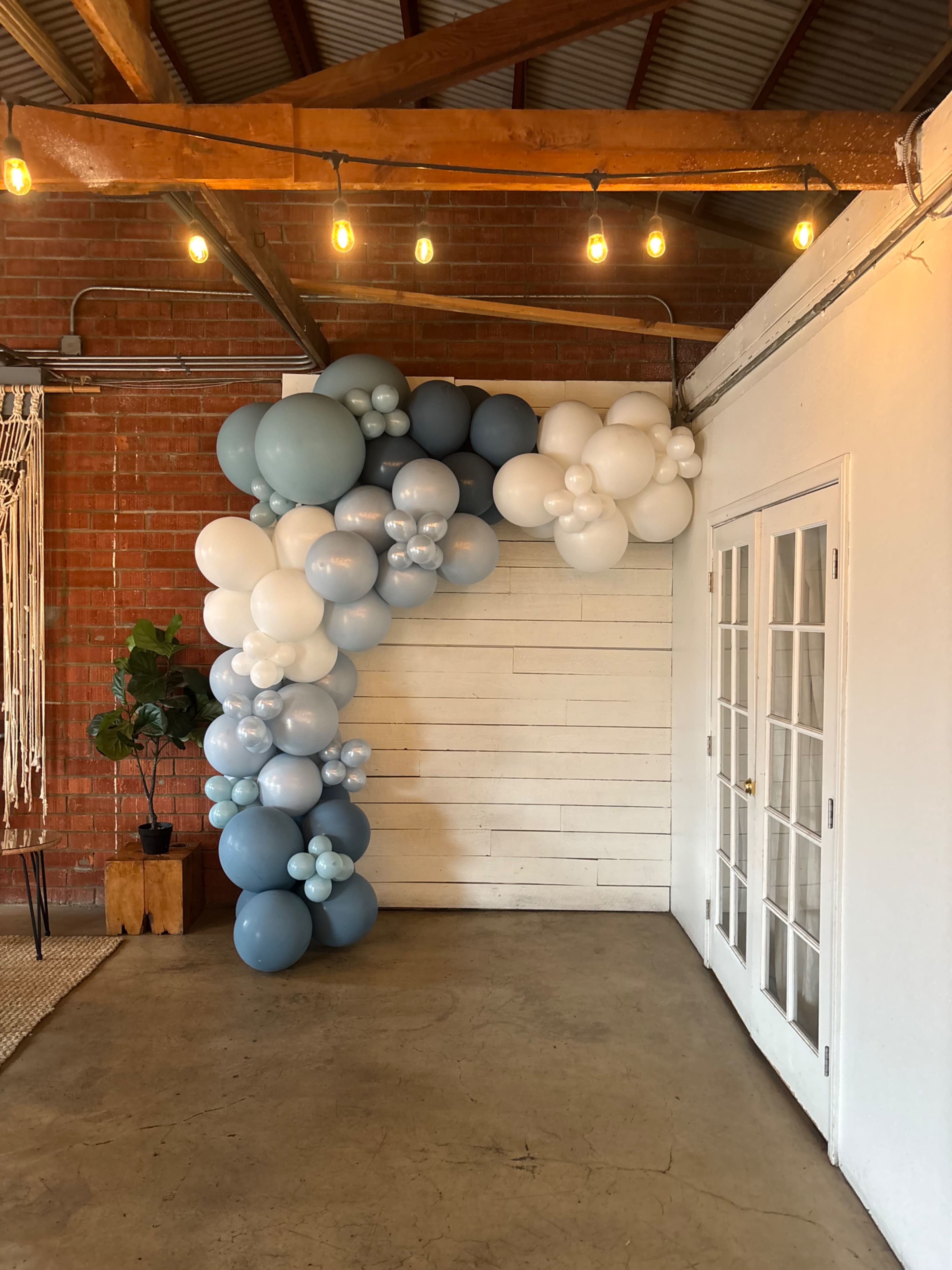 A cluster of balloons in shades of blue and white forms an archway against a white wooden wall in a room with exposed brick and wooden beams.
