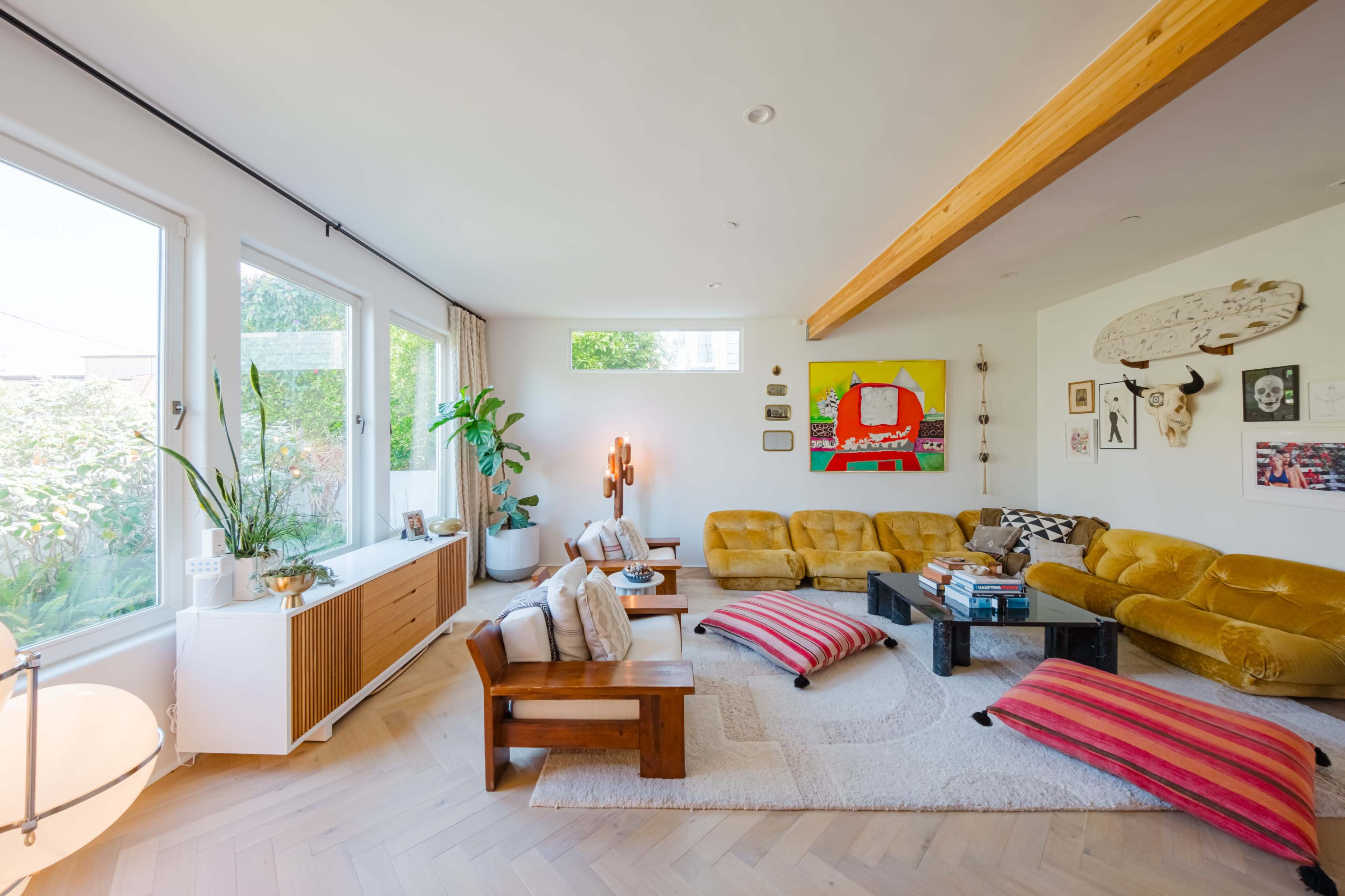 A spacious living room with large windows, a mix of seating options in yellow and neutral tones, and decorative artwork on the walls.