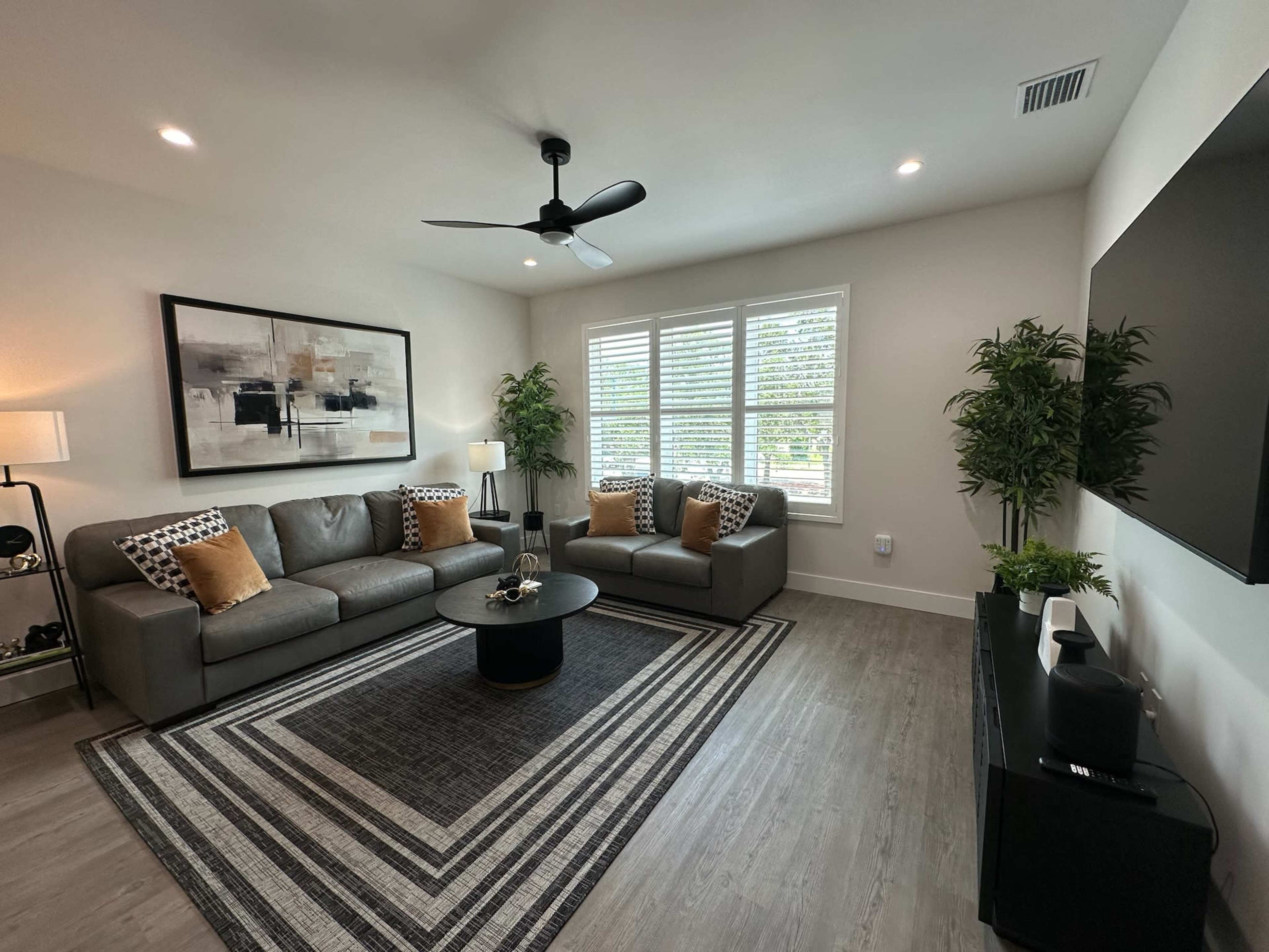 A modern living room with a gray sectional couch, a round black coffee table, and a large wall-mounted TV, surrounded by decorative plants and a geometric area rug.
