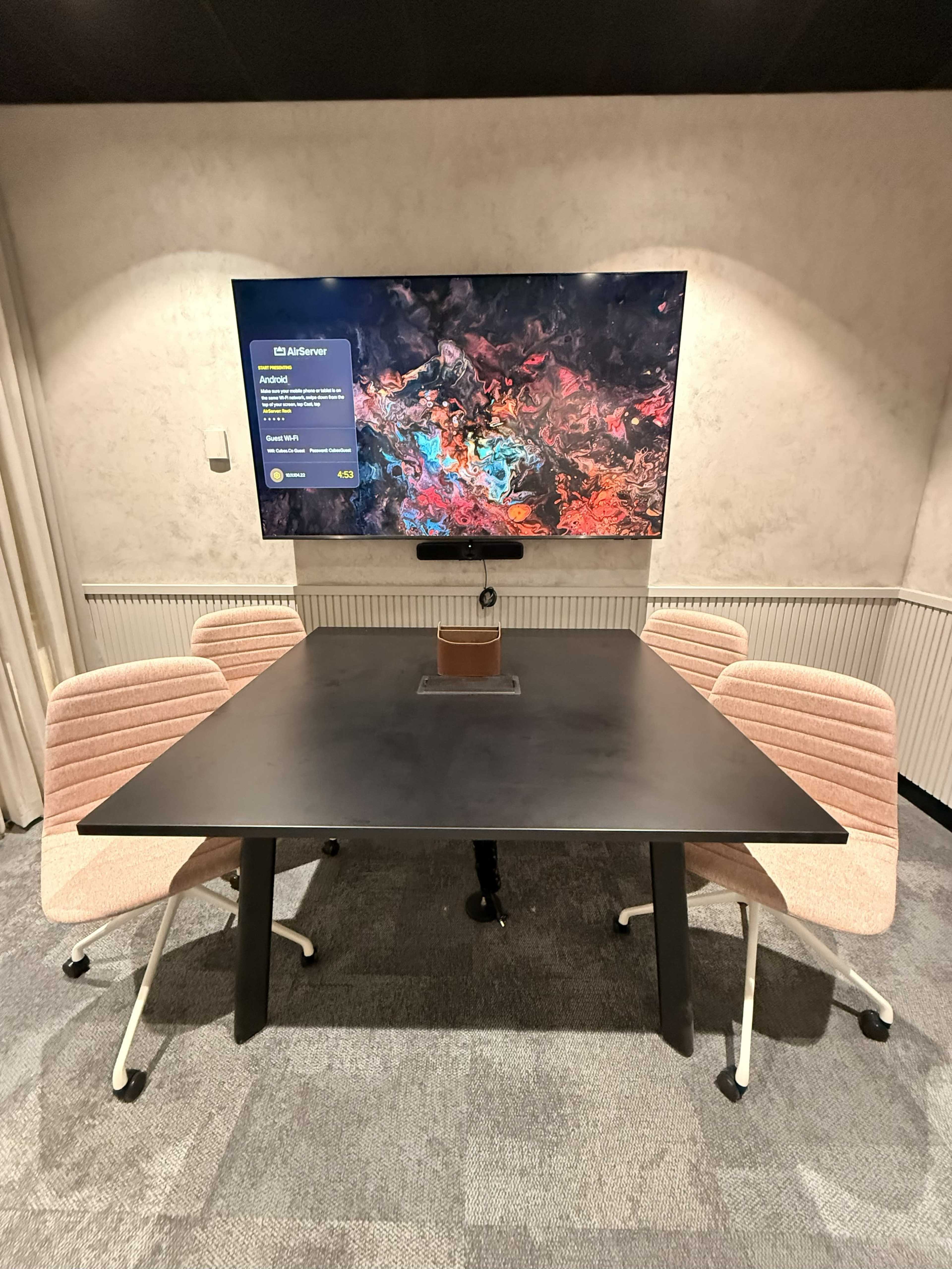 A modern conference room features a black table surrounded by pink chairs and a large screen displaying vibrant abstract art on the wall.