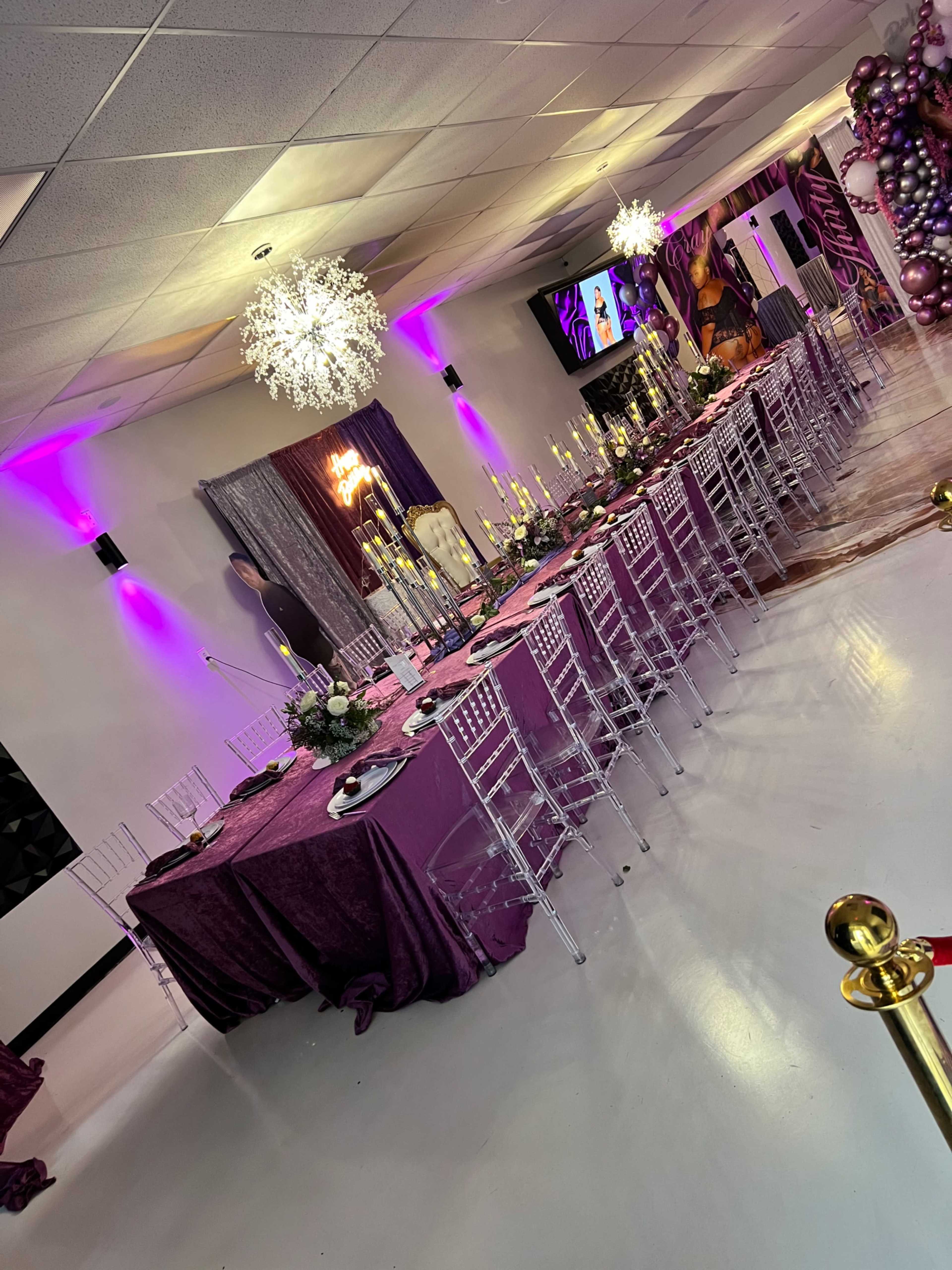 A large dining area set up with a long table covered in purple linens, surrounded by clear chairs and adorned with floral arrangements and candles, under illuminated chandeliers.