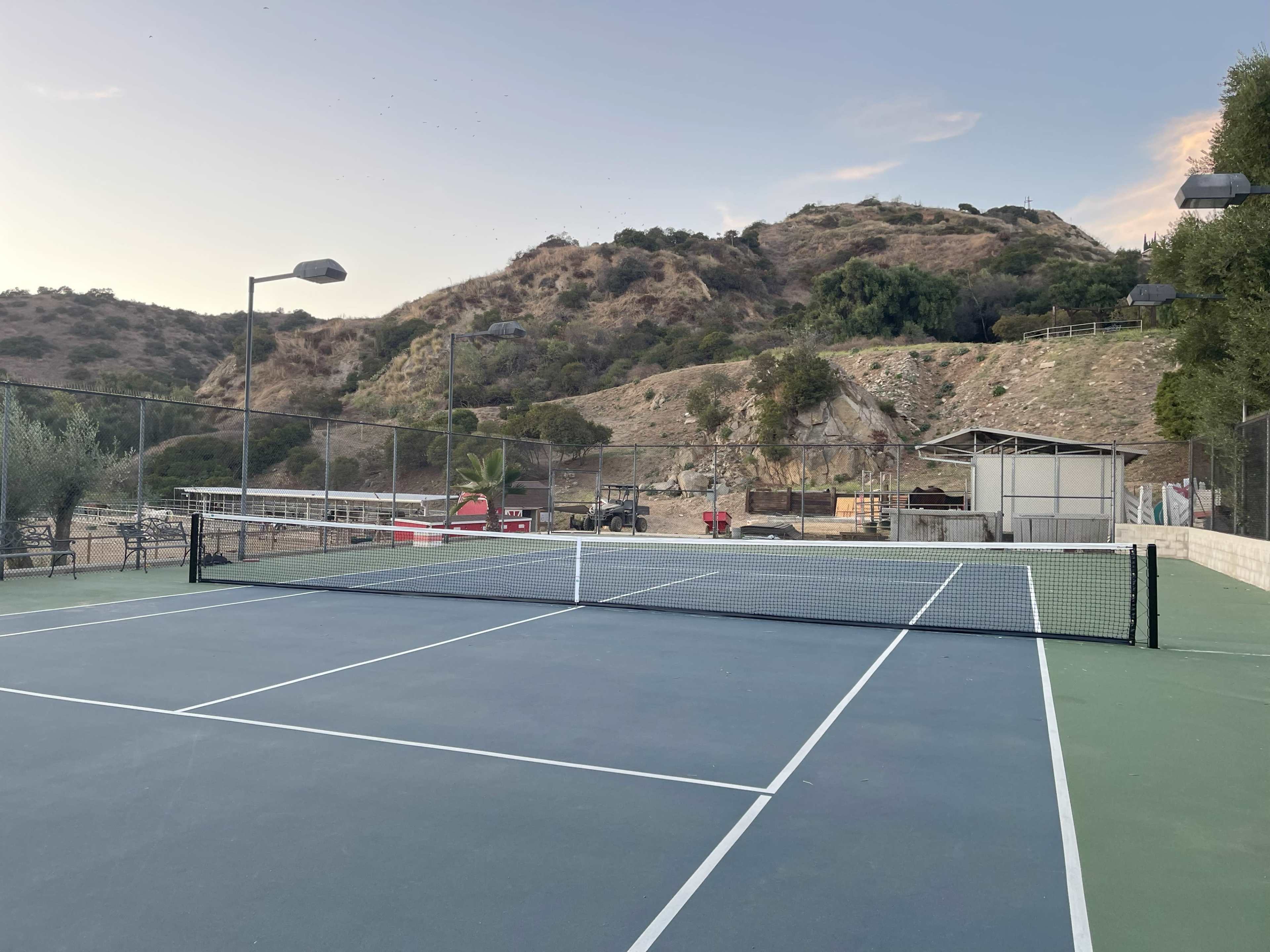 A tennis court is set against a backdrop of rolling hills and trees, with a nearby building and lights for evening play.