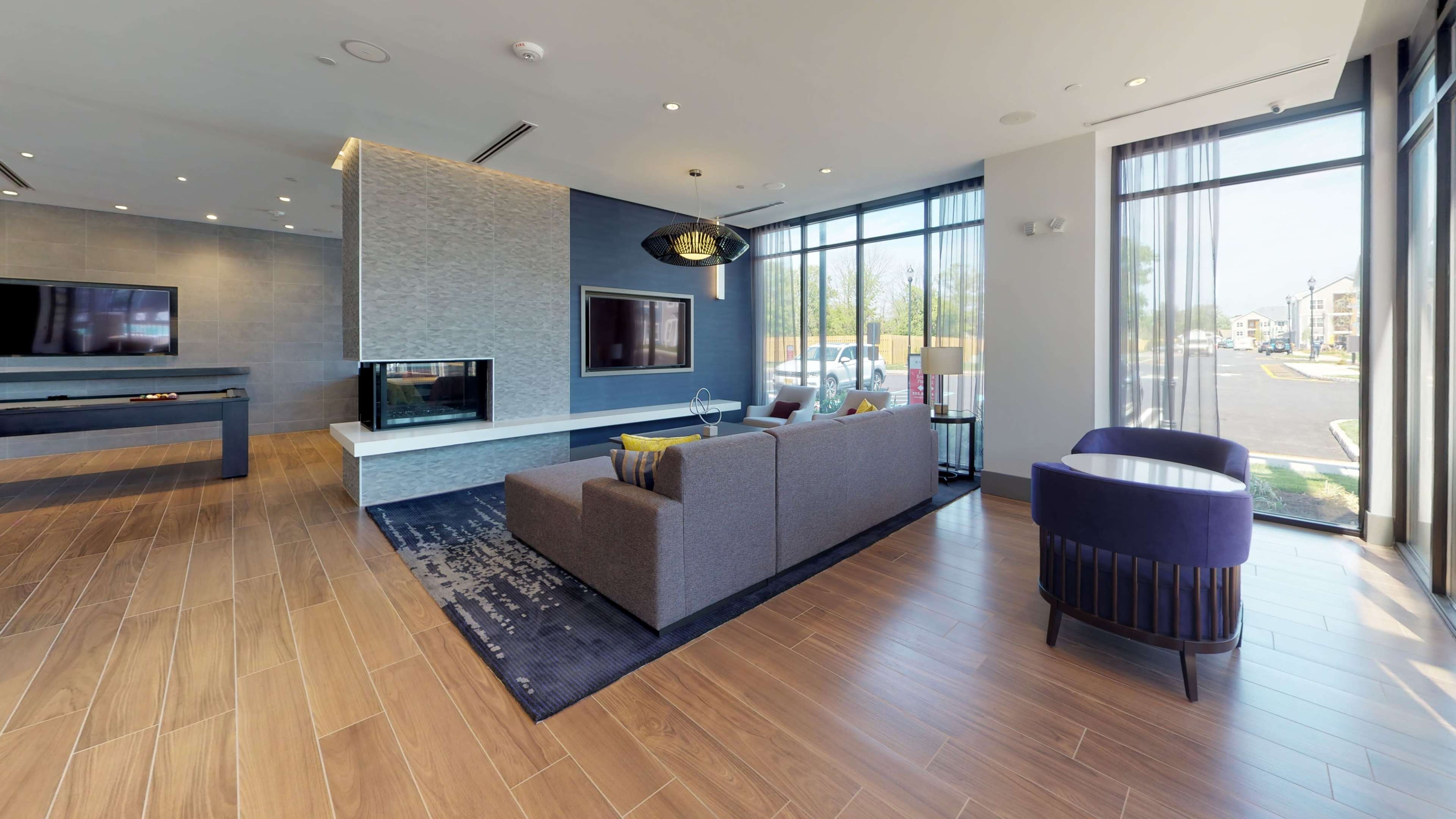 A modern lounge area featuring a gray sofa, a fireplace, television screens, and large windows overlooking an outdoor space.