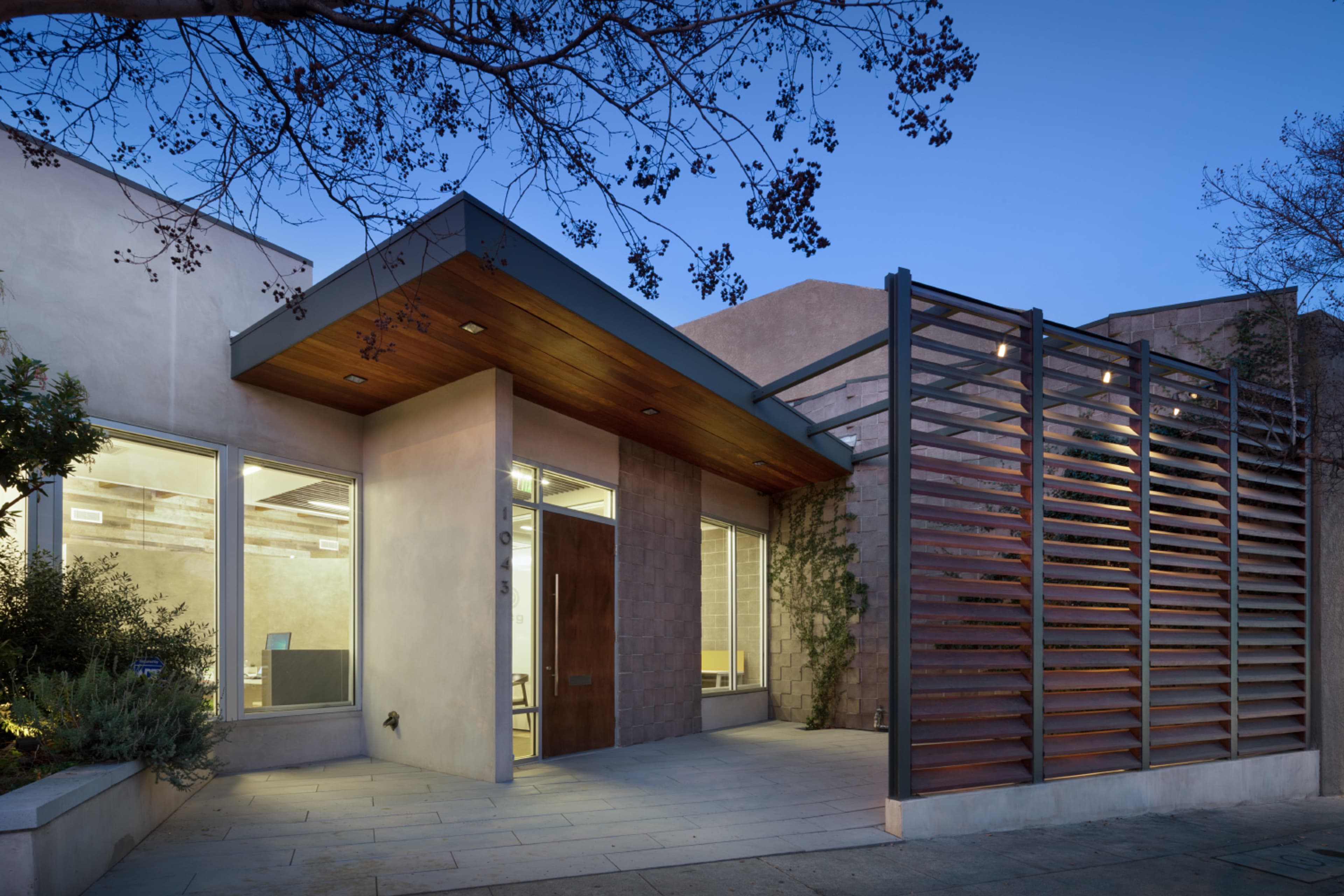 A modern, single-story building with a wooden overhang, large windows, and a slatted wooden screen at the entrance, set against a twilight sky.