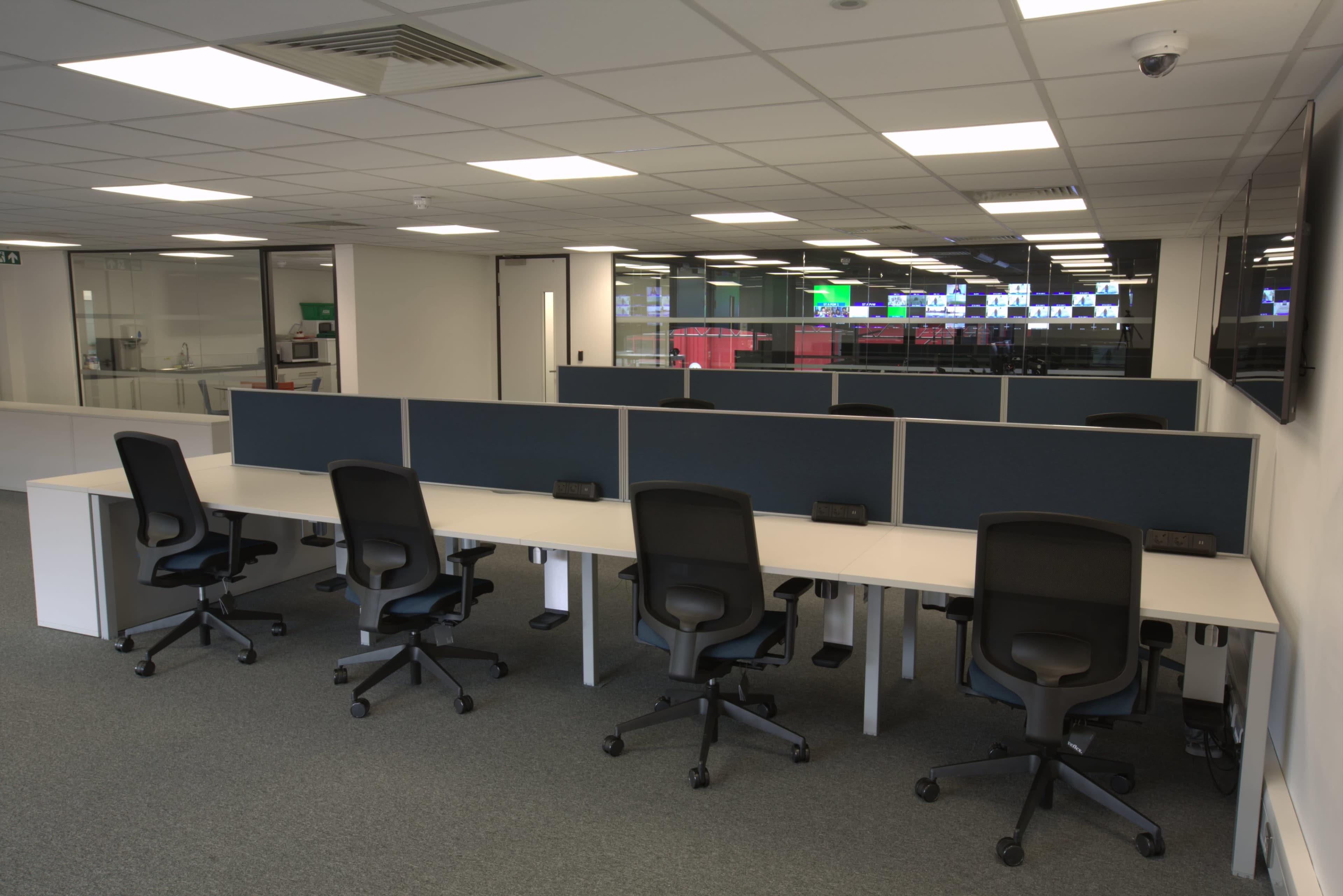 The image shows a modern office space with several empty workstations equipped with black rolling chairs and partitioned desks.
