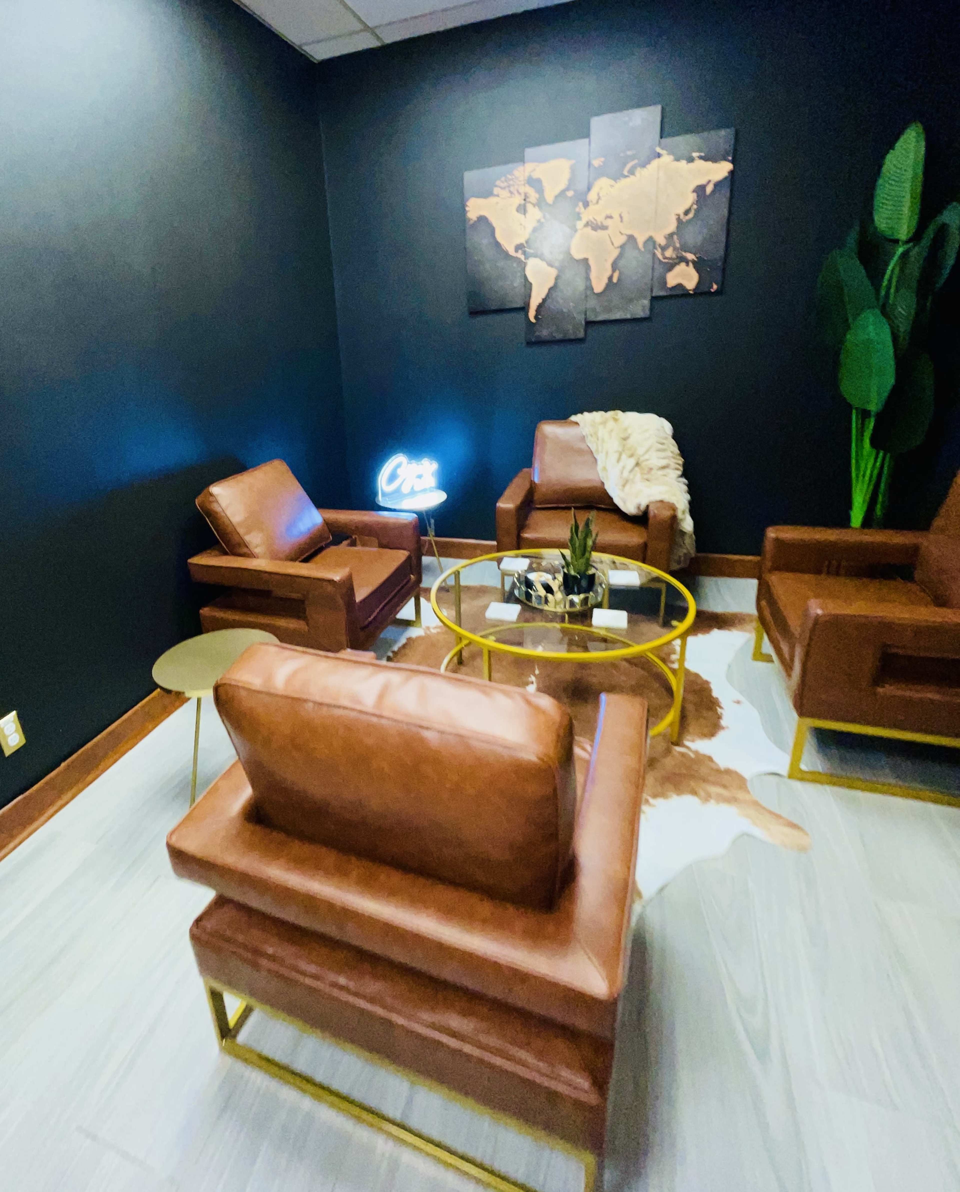A small lounge area featuring two brown leather chairs, a round coffee table with decorative items, a cowhide rug, and a world map on the wall, all complemented by a neon sign.
