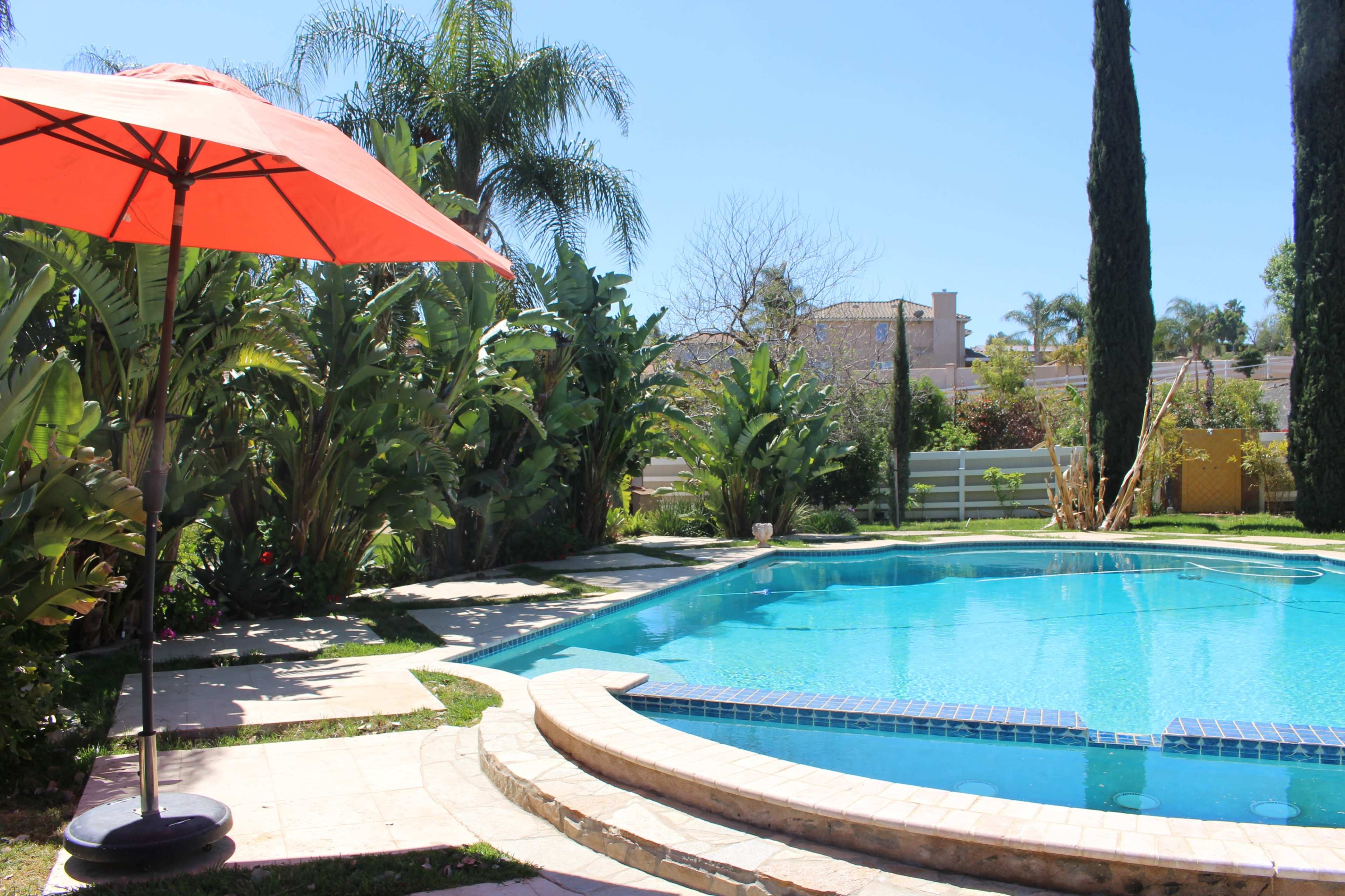 A bright orange umbrella shades a lounge area near a clear blue swimming pool surrounded by lush greenery.