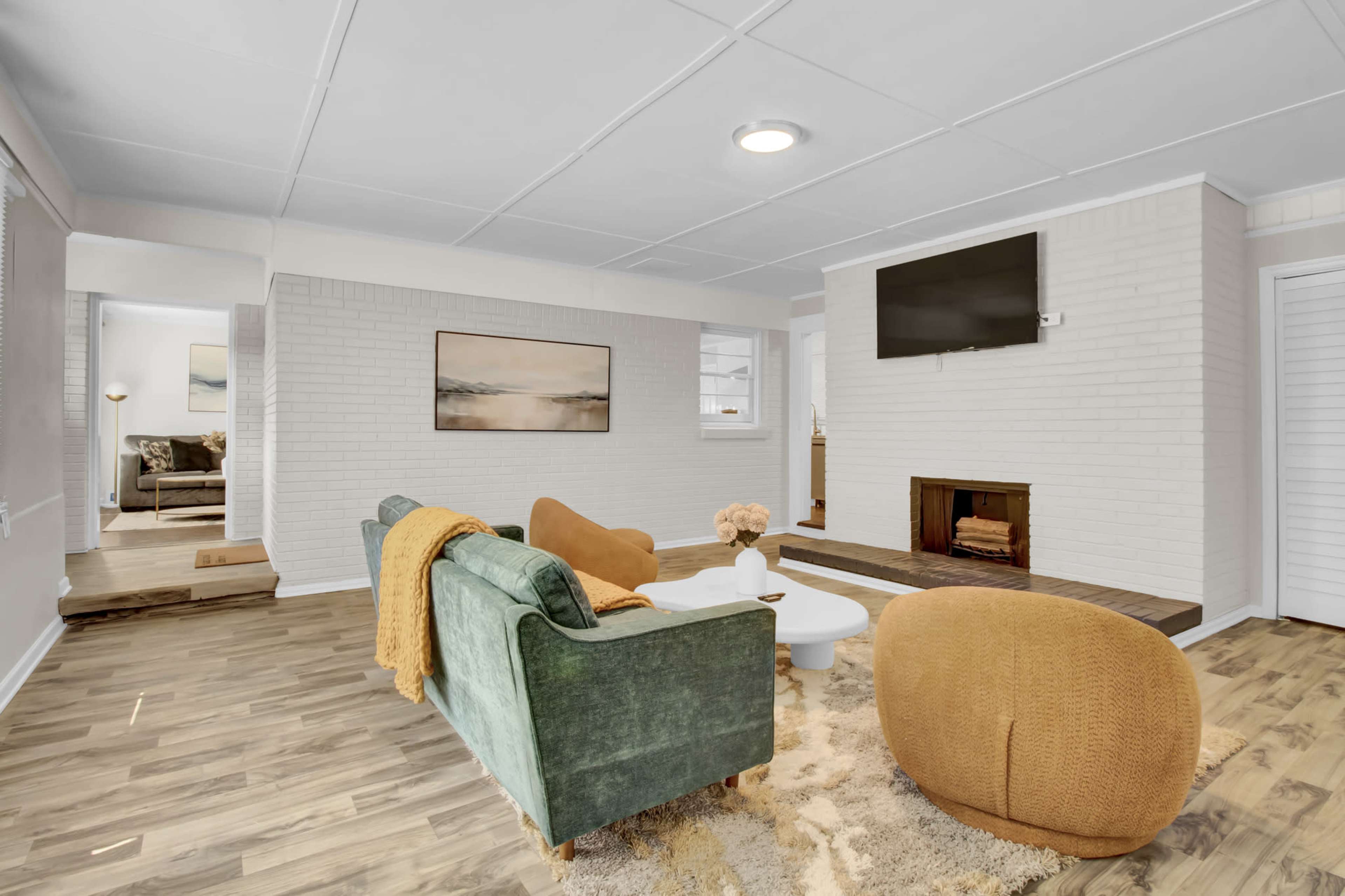 The living room features a green sofa, round brown chairs, a white coffee table, and a wall-mounted television above a brick fireplace.
