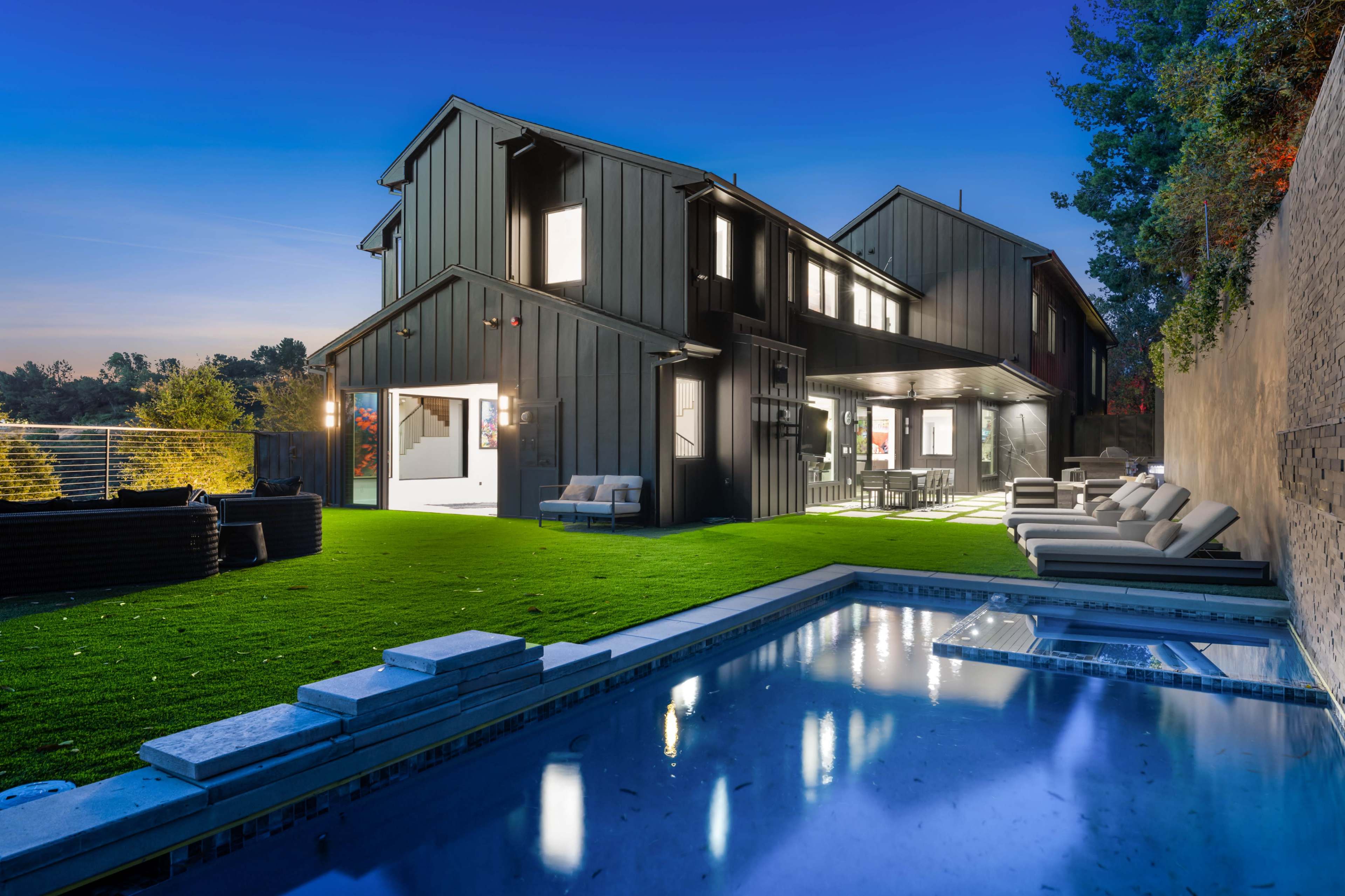 The image shows a modern two-story black house with a swimming pool and lounge chairs in a landscaped backyard during twilight.