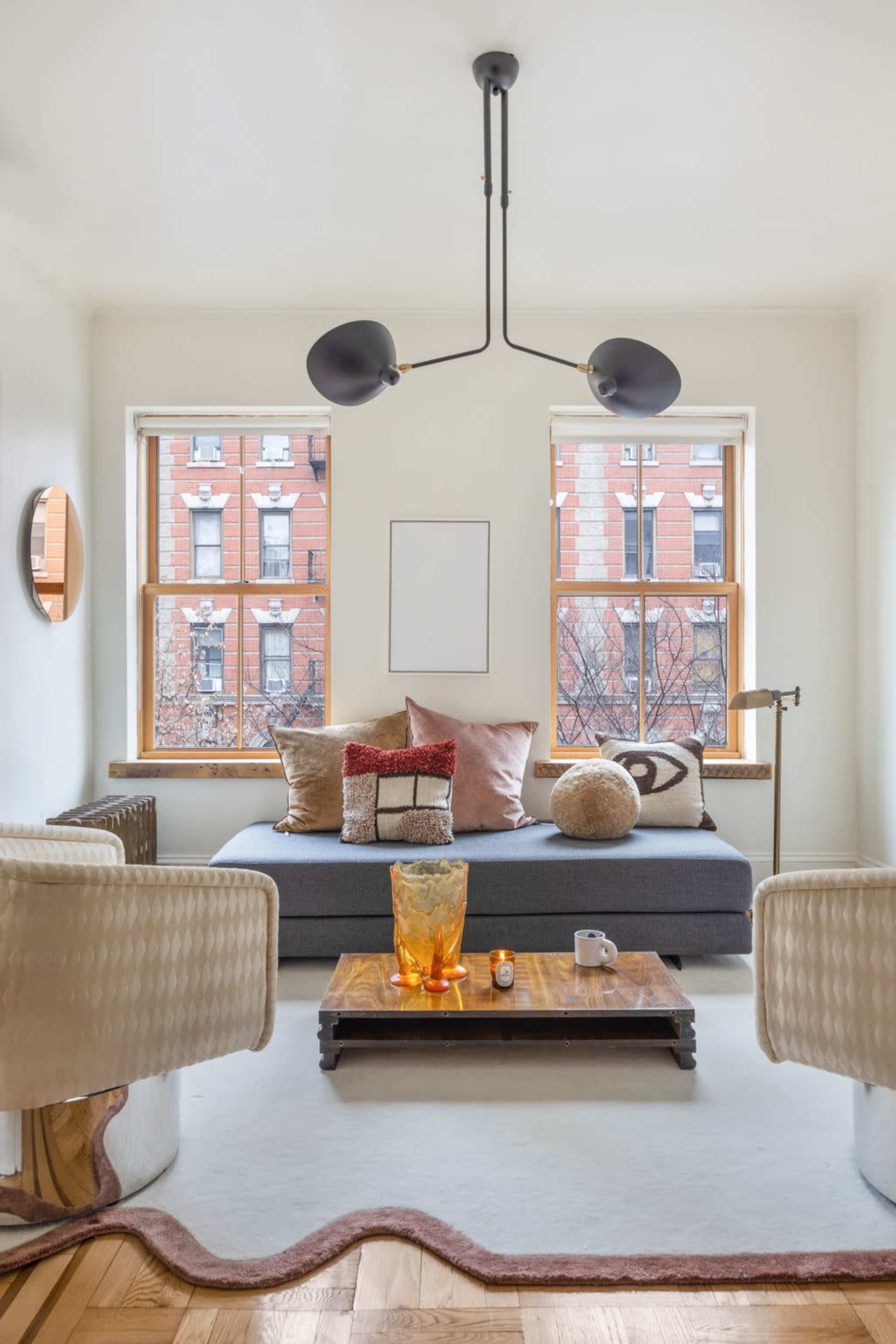 A cozy living room features a gray sofa with decorative pillows, a wooden coffee table, and large windows displaying a brick building outside.