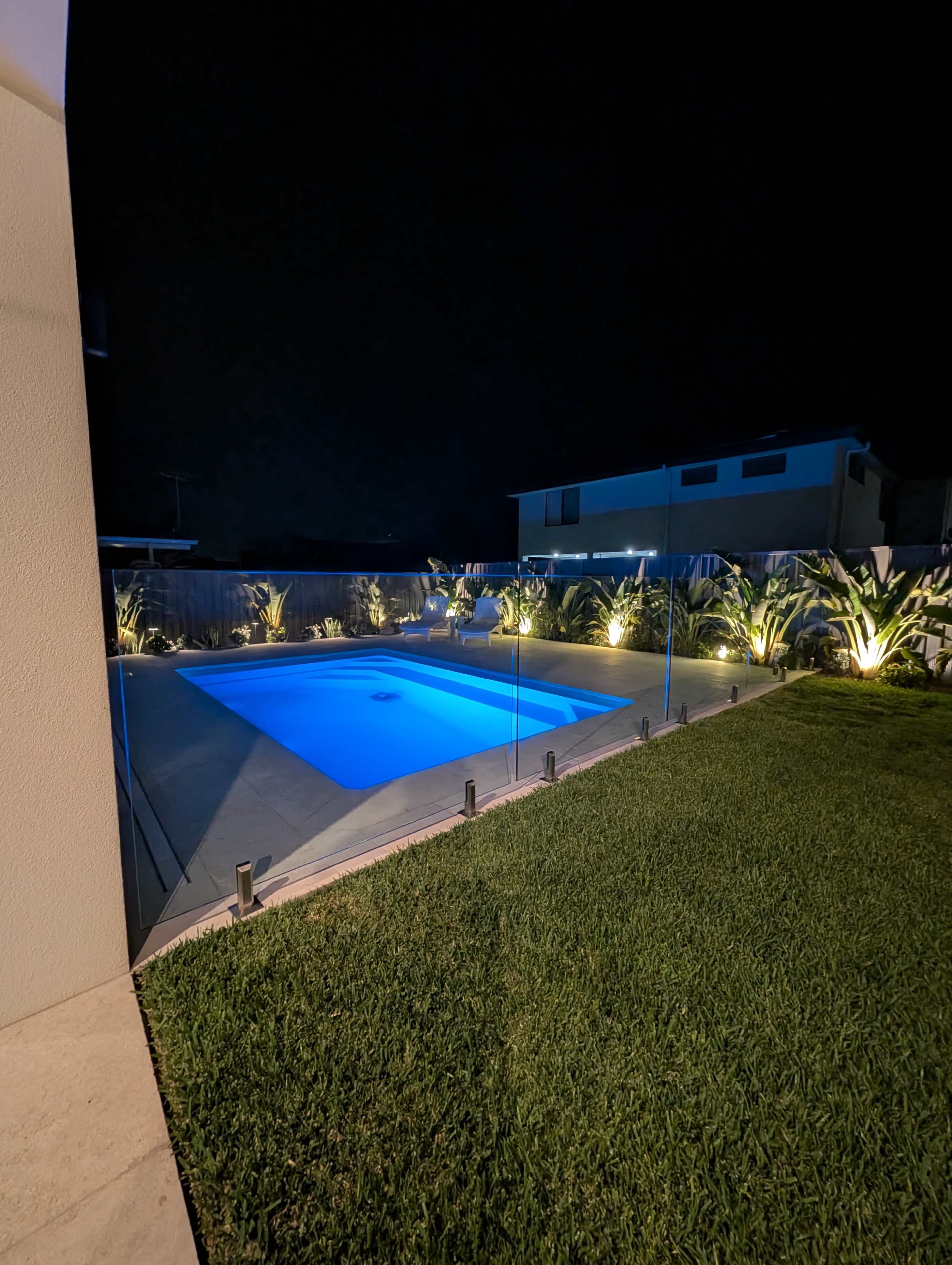 A well-lit swimming pool with blue water is surrounded by grassy areas and illuminated landscaping at night.