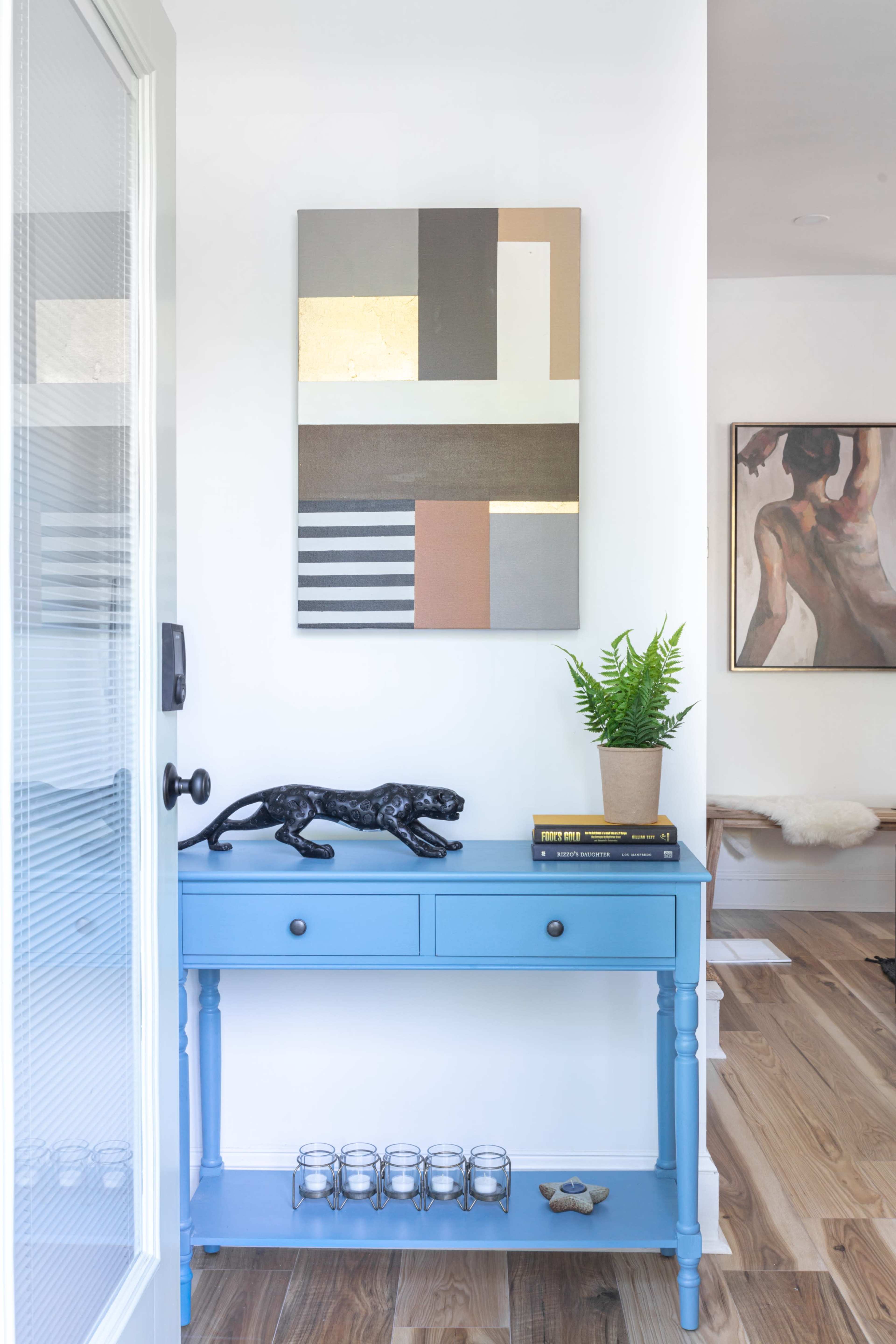 A blue console table with a black panther statue and a potted fern sits against a wall adorned with abstract art, with hardwood flooring visible in the background.