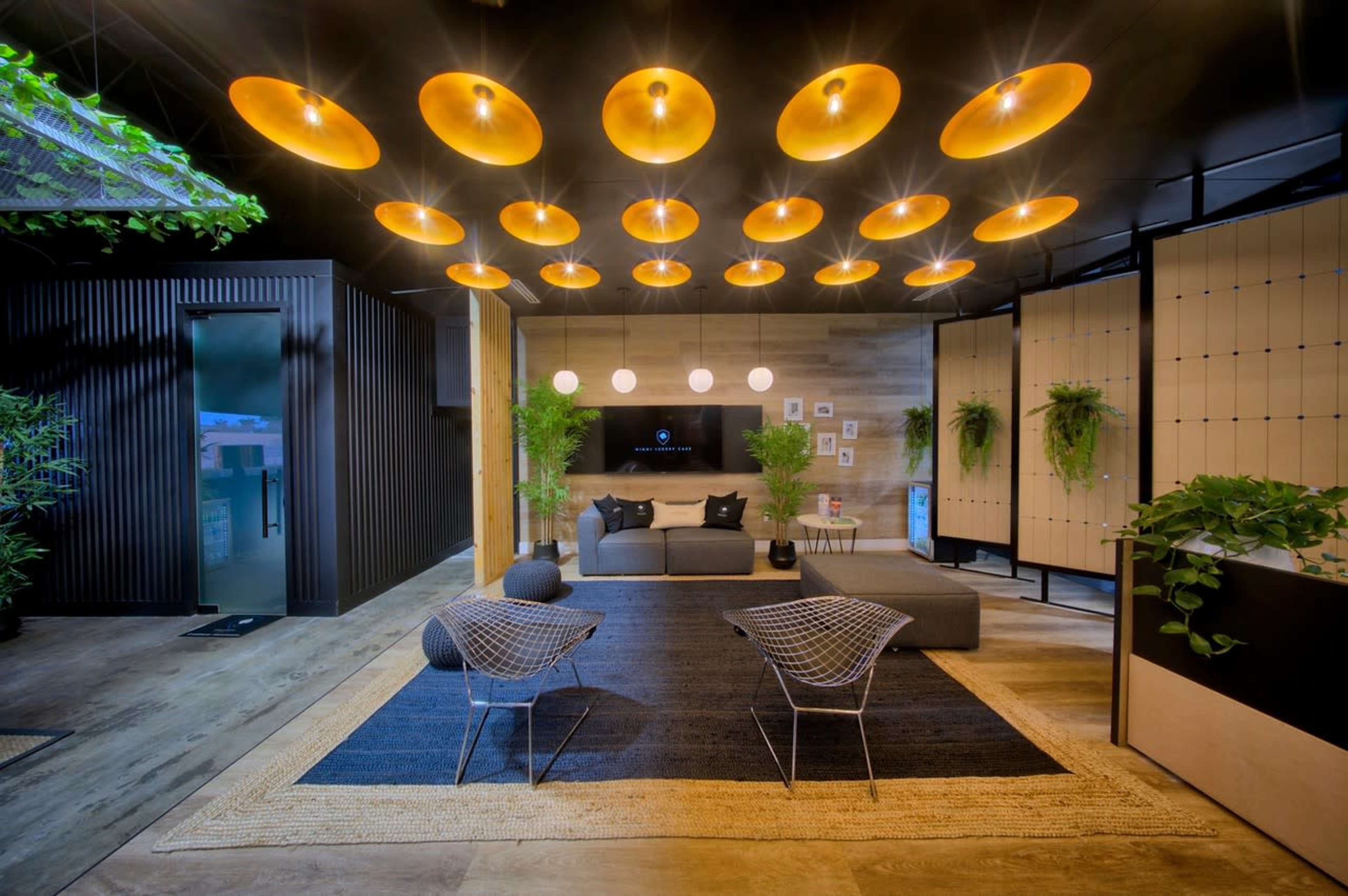 A modern living room features a gray sofa, geometric chairs, and overhead pendant lights, surrounded by wall plants and a television.
