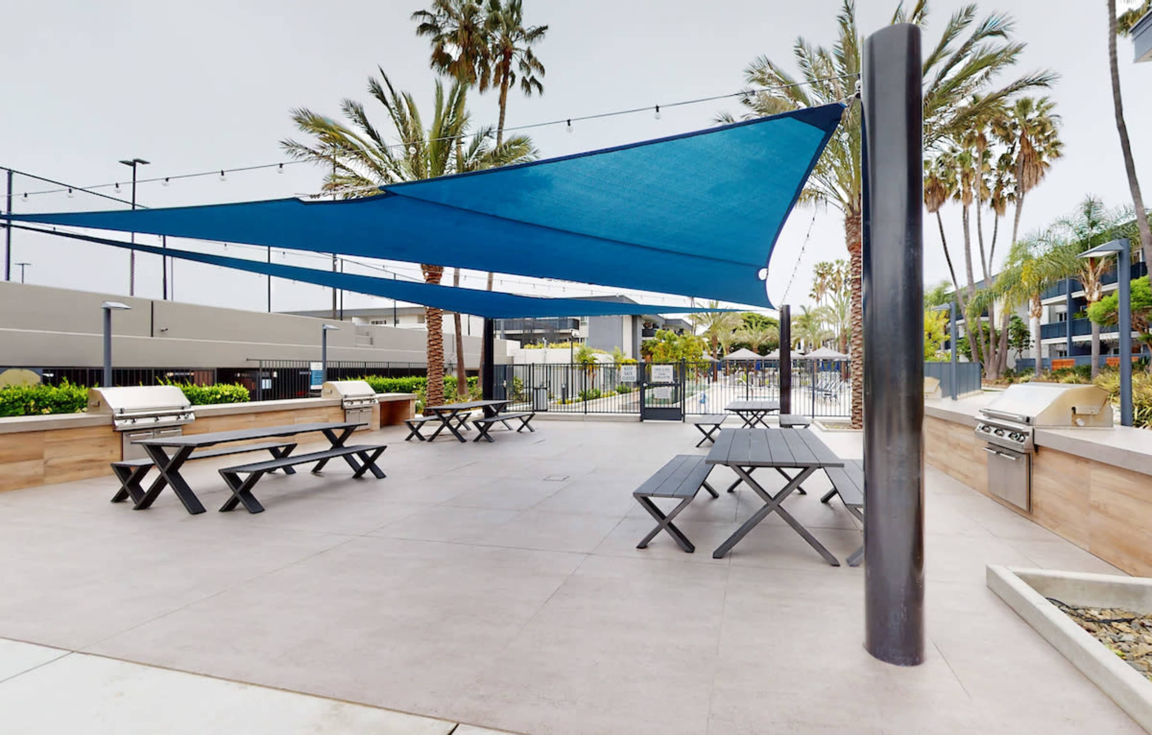 An outdoor barbecue area with several picnic tables shaded by blue canopies, surrounded by palm trees.