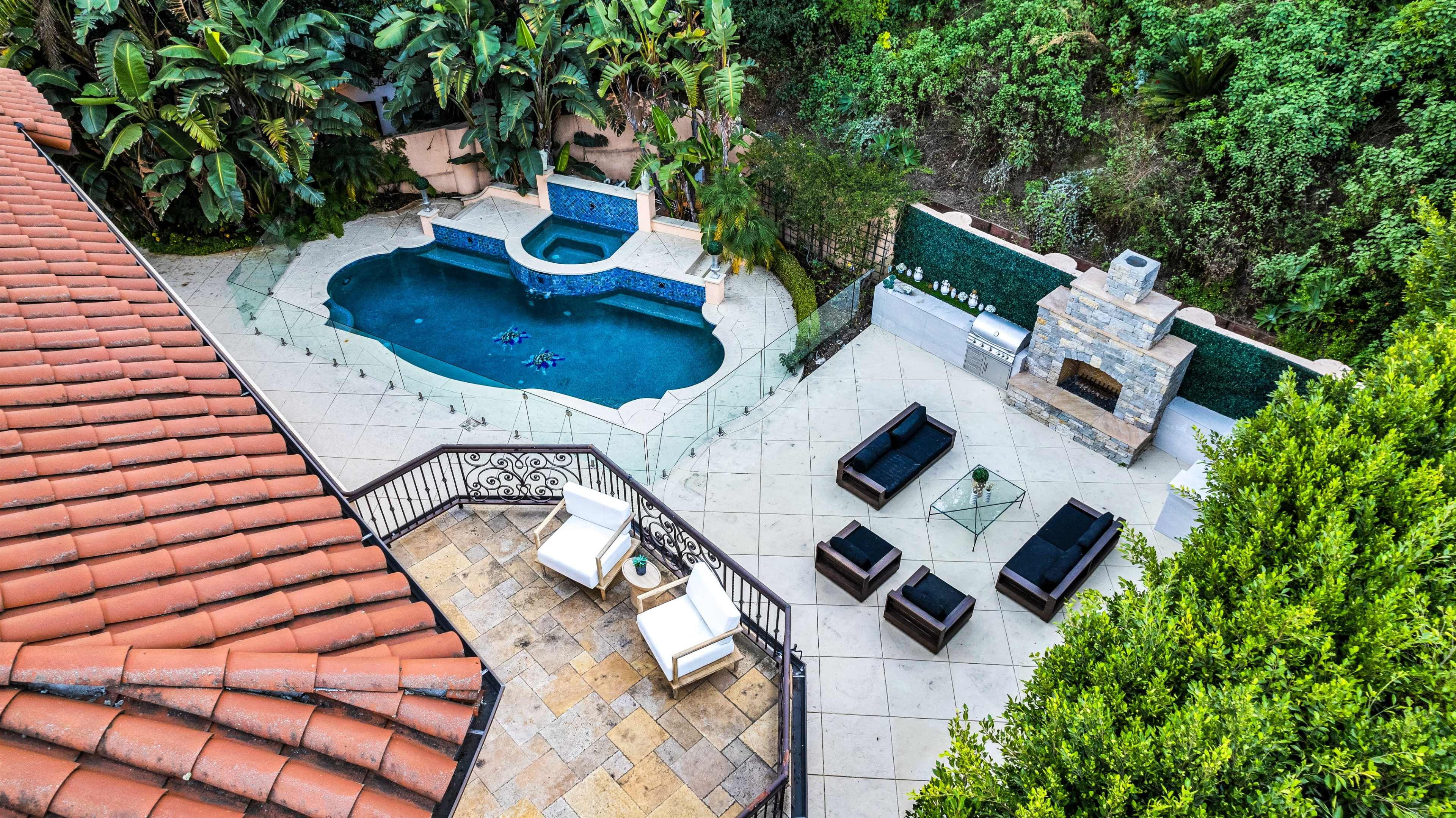 The image shows a backyard featuring a swimming pool surrounded by lounge chairs and a stone fireplace, with lush greenery in the background.