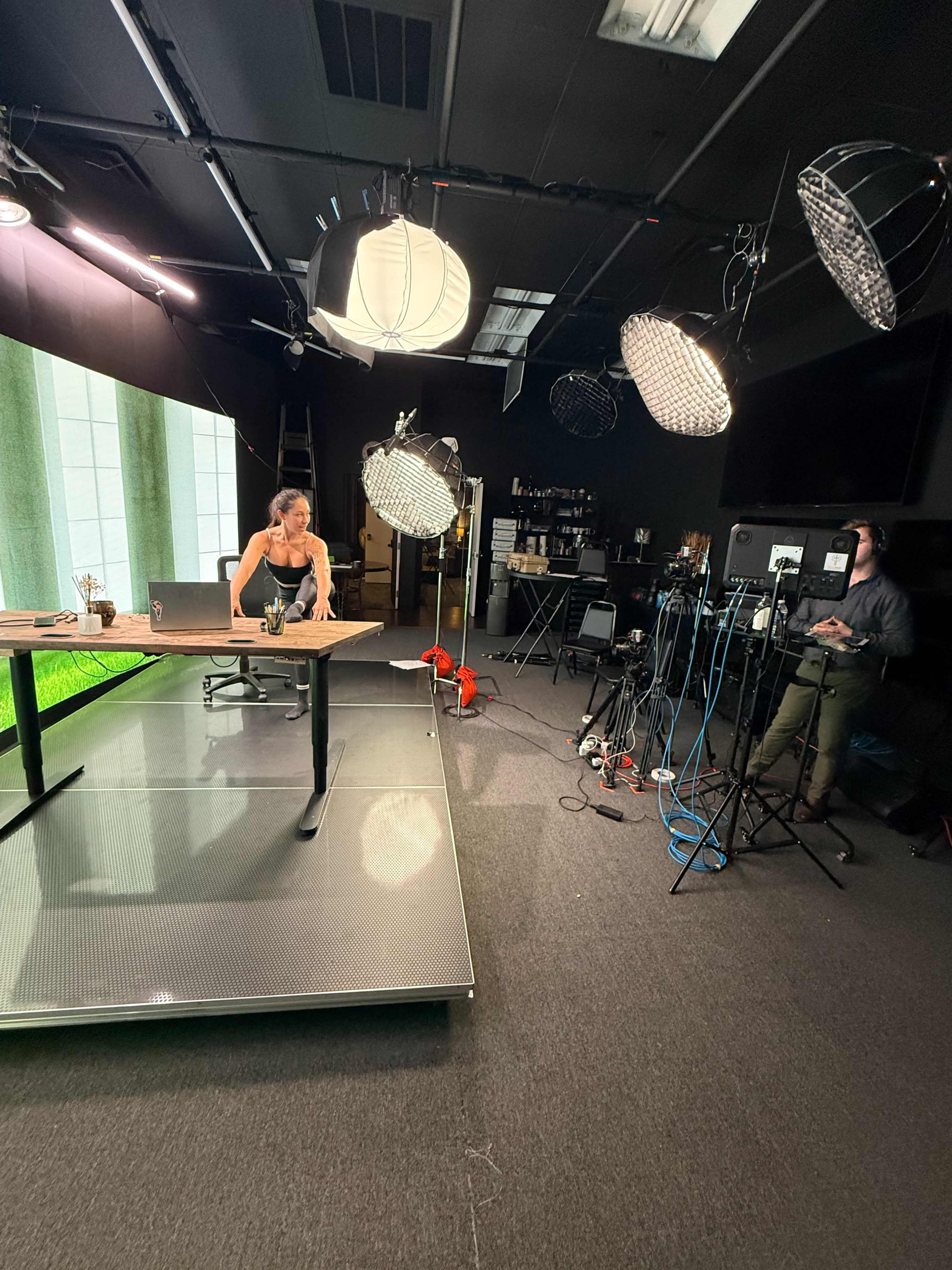 A woman is working at a table in a well-lit studio setting equipped with multiple lighting sources and cameras.