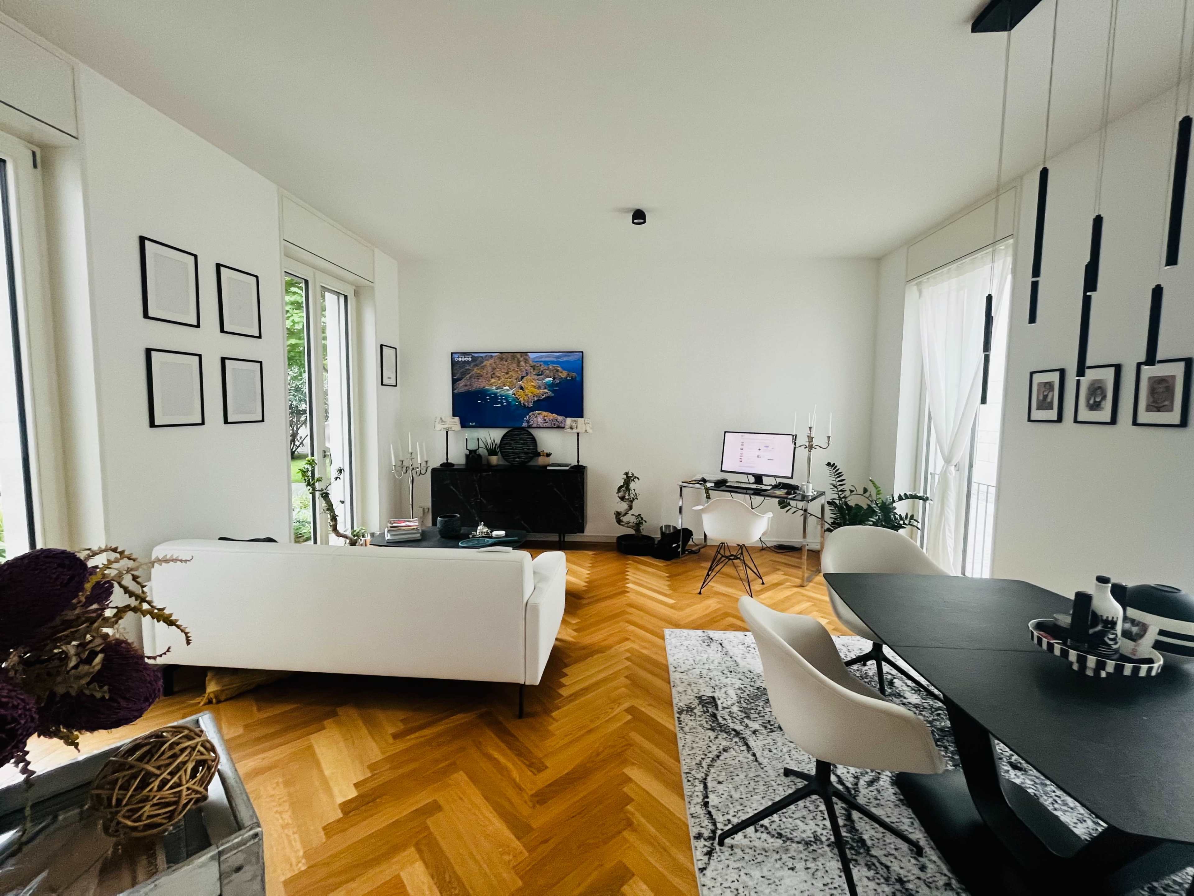 A modern living room with a white sofa, a black dining table, and large windows allowing natural light to enter.