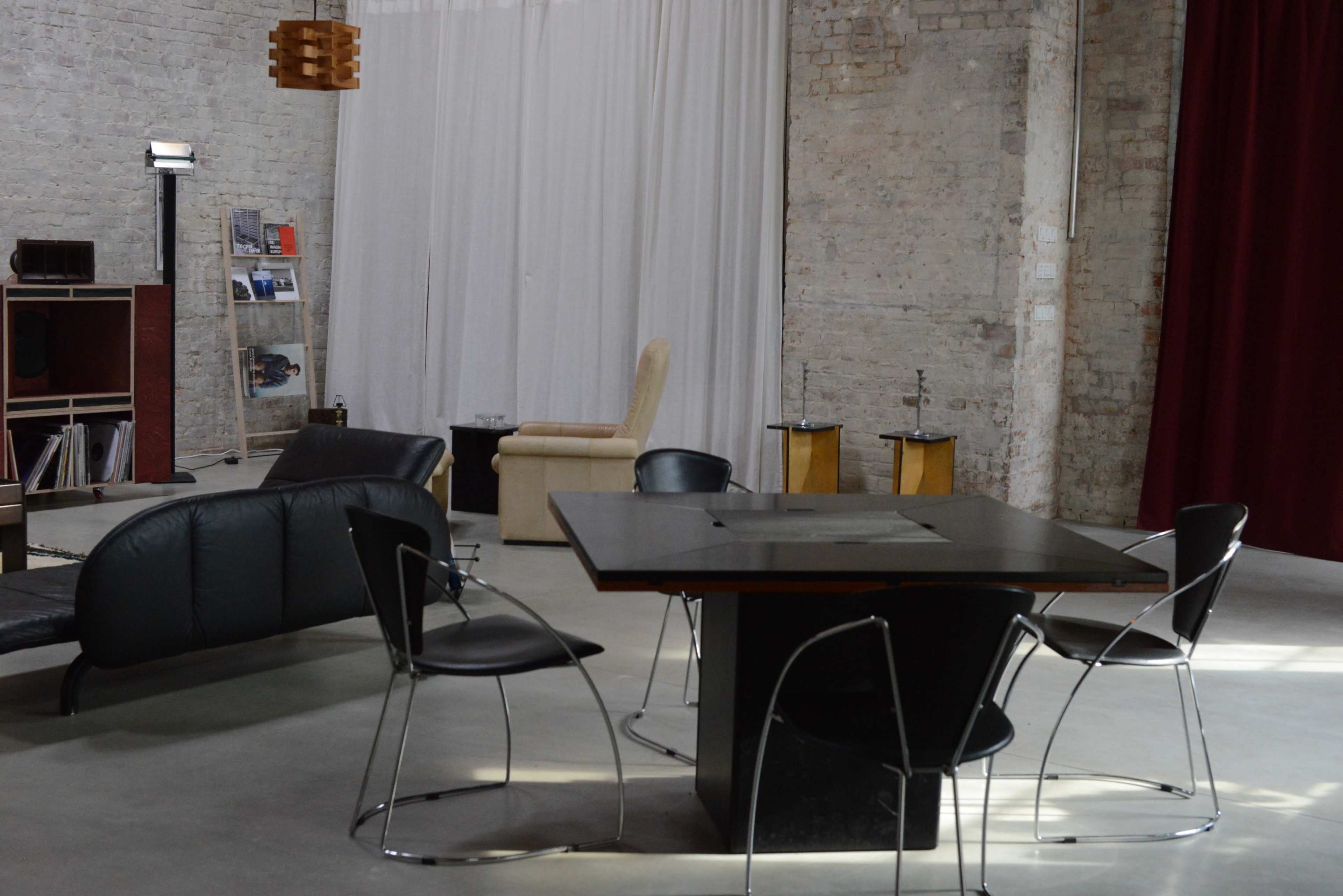The image shows a modern interior space featuring a black table surrounded by four chairs, a lounge area with a leather sofa, and a bookshelf against a brick wall.