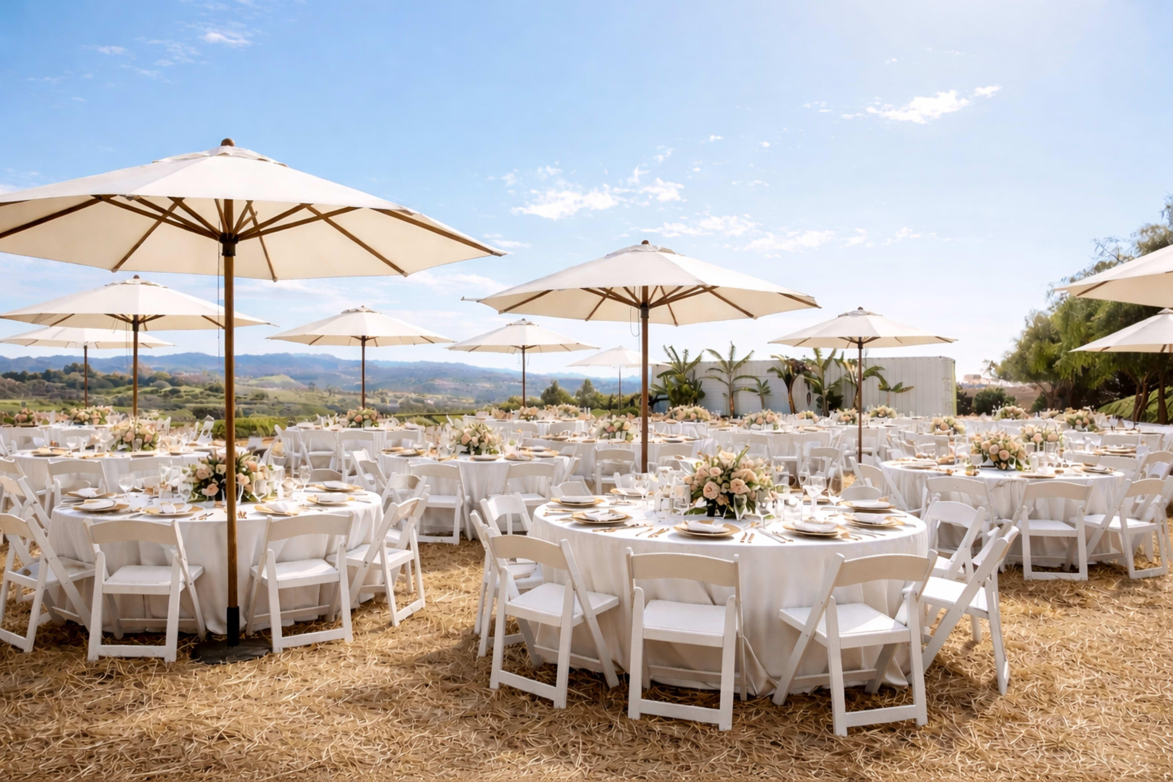 A beautifully arranged outdoor wedding reception features round tables adorned with floral centerpieces under large white umbrellas, set against a scenic landscape.