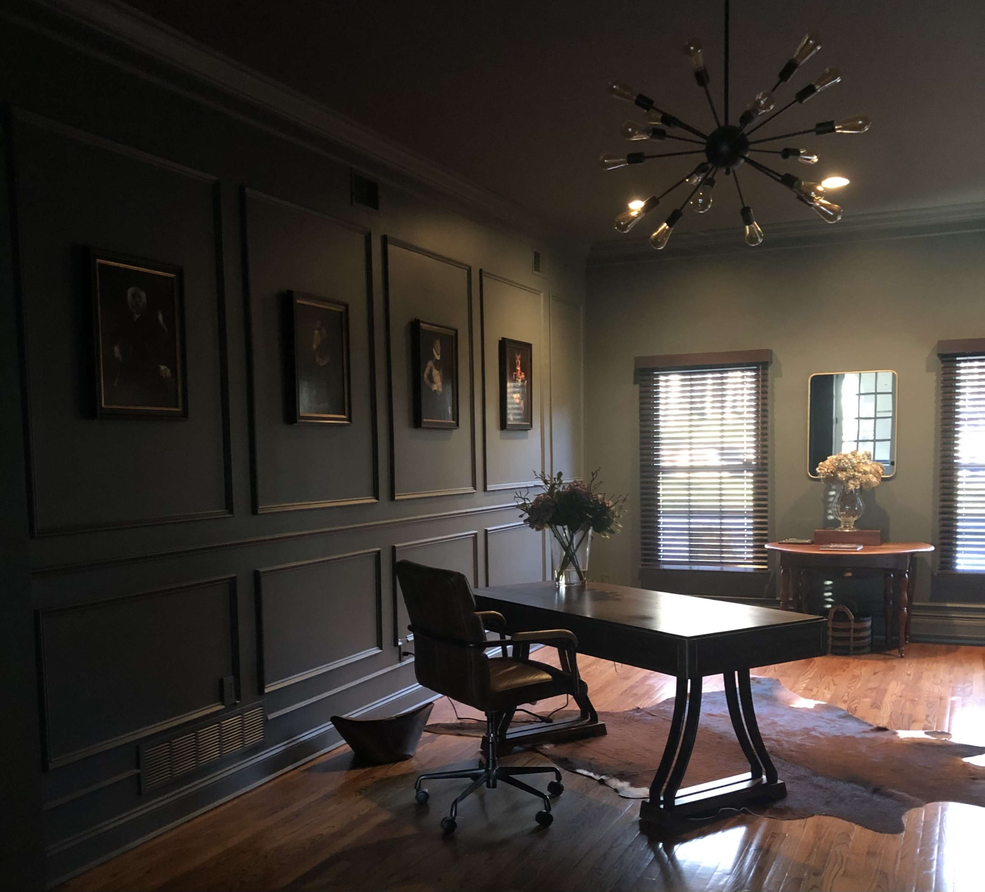 The image shows a dimly lit office space with a wooden desk, an office chair, and framed artwork on the walls, featuring a large mirror and a vase of flowers on a nearby table.