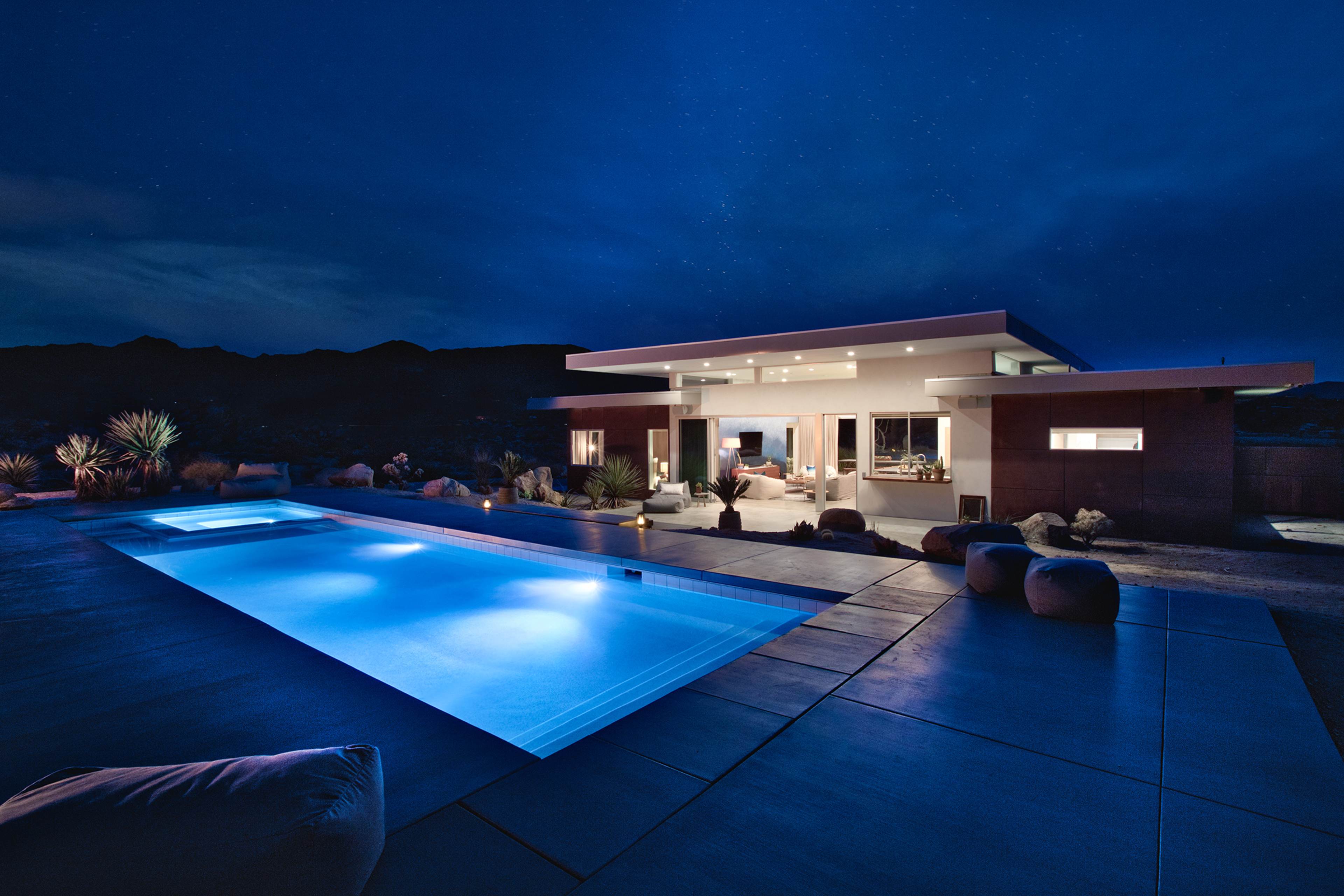 A modern house with a swimming pool is illuminated at night, set against a starry sky in a desert landscape.
