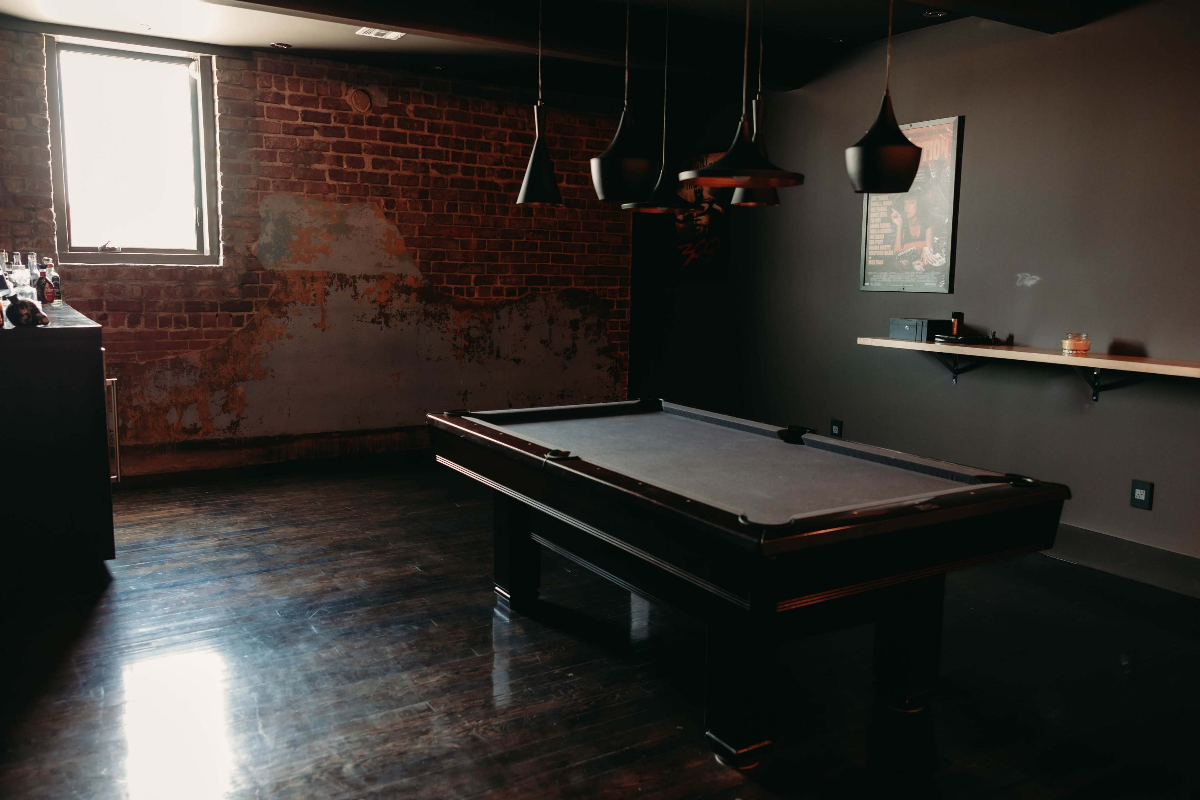 A billiard table is centered in a dimly lit room with exposed brick walls and a minimalist bar in the background.