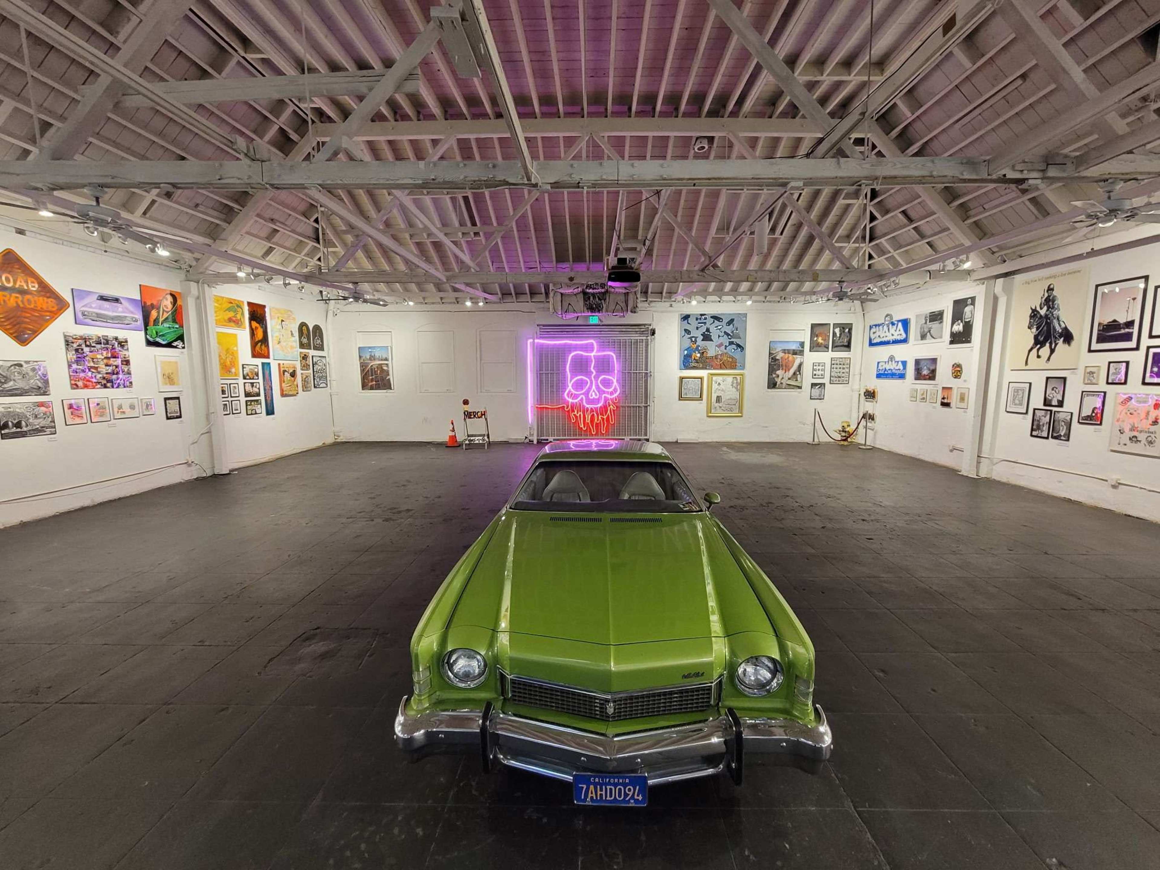 A green car is positioned in the center of an art gallery, surrounded by various framed artworks on the walls.