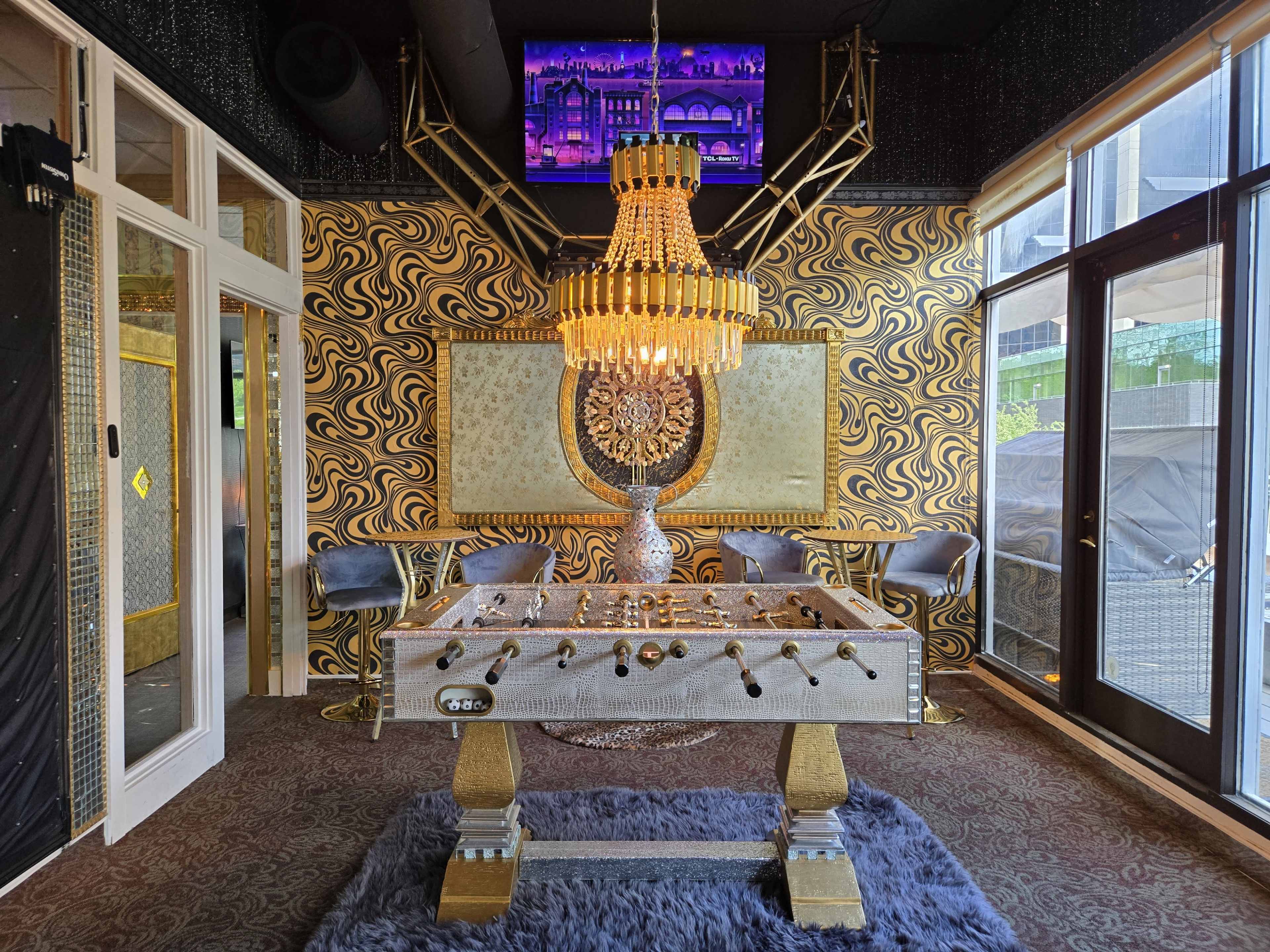 The image shows a stylish game room featuring a foosball table, a large chandelier, and a patterned wall with a television mounted above.
