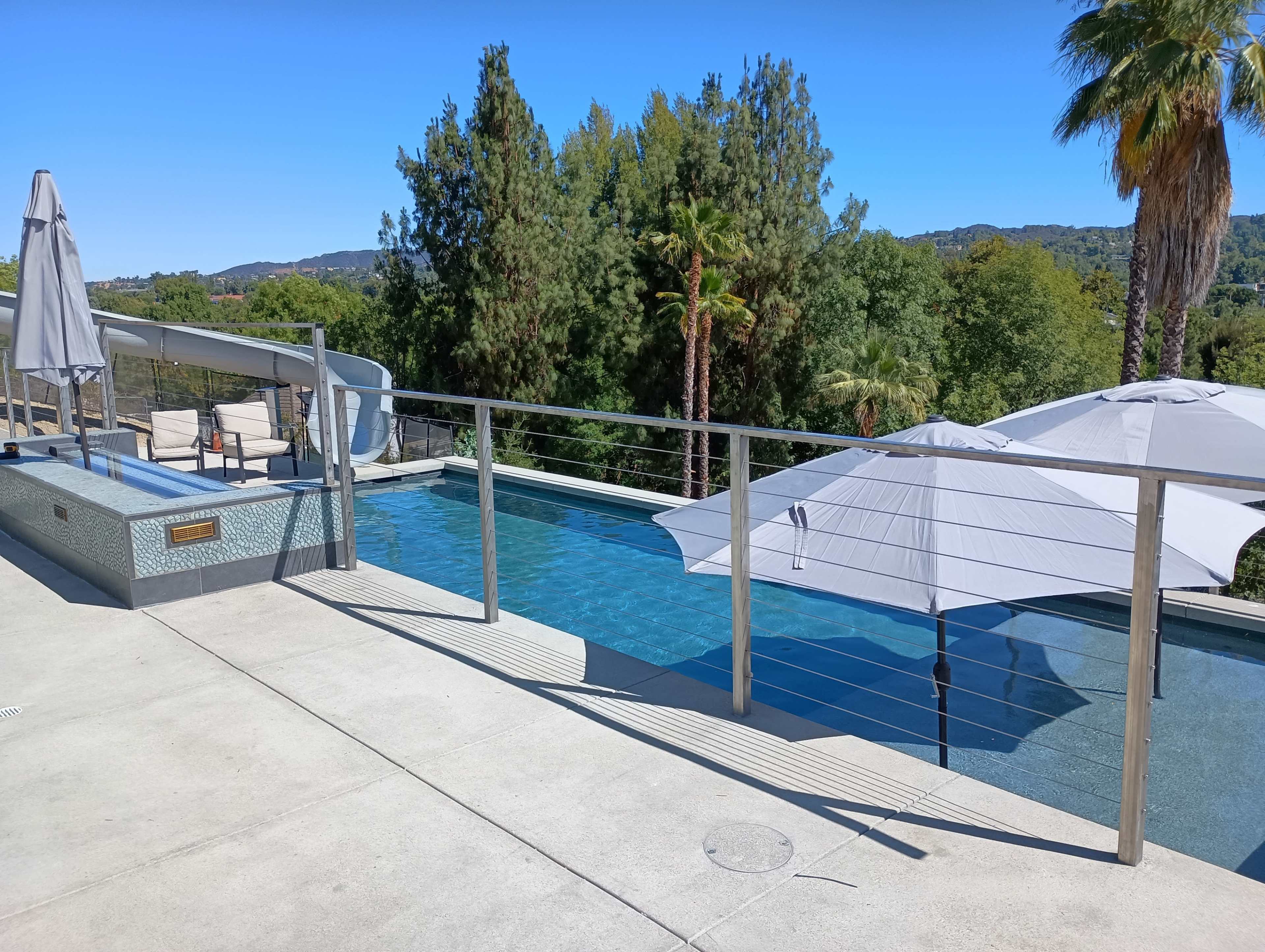 The image shows a modern swimming pool area with a slide, two large umbrellas, and lush greenery in the background.