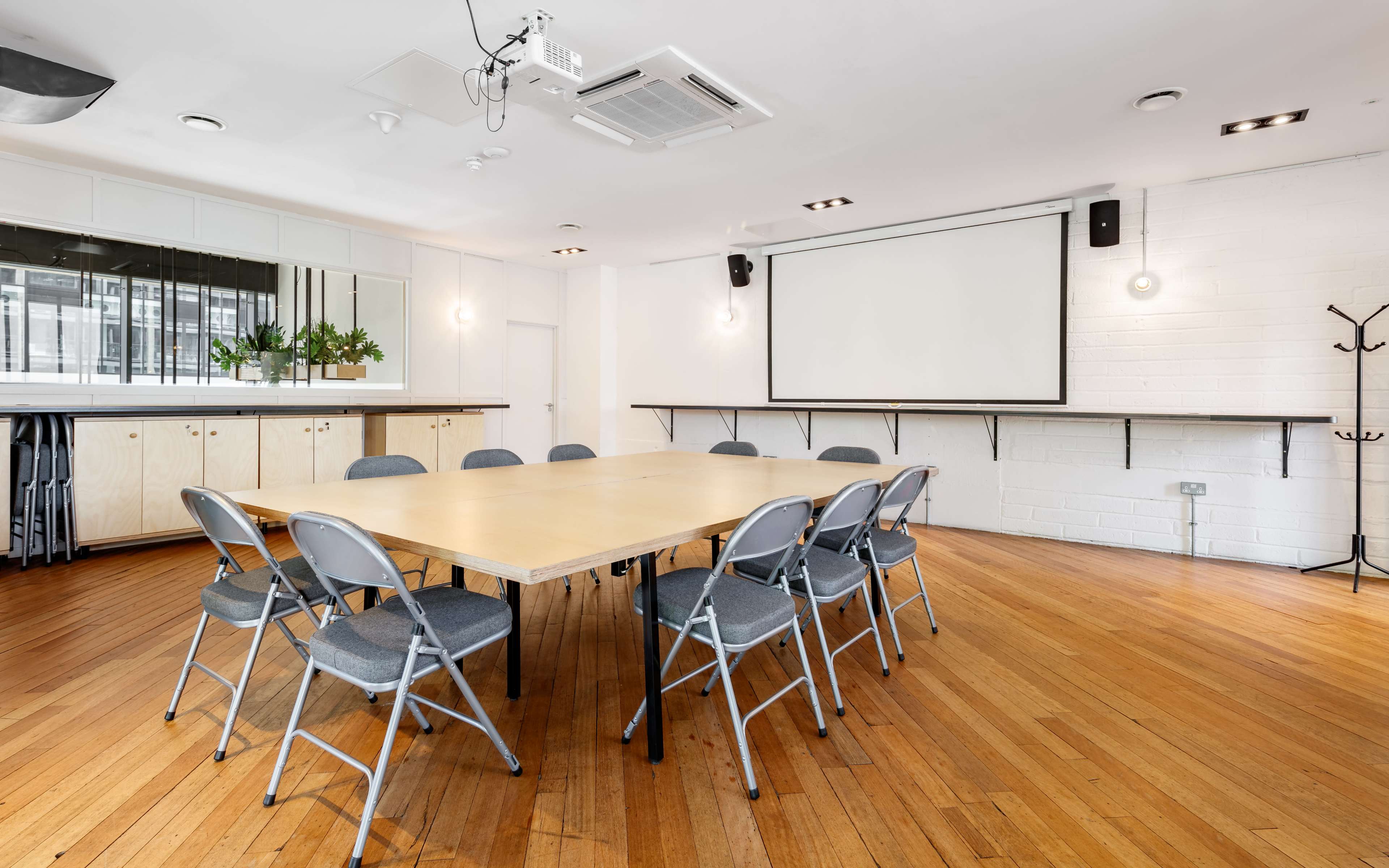 A conference room features a rectangular table surrounded by eight folding chairs, with a wall-mounted screen and a wooden bar area in the background.
