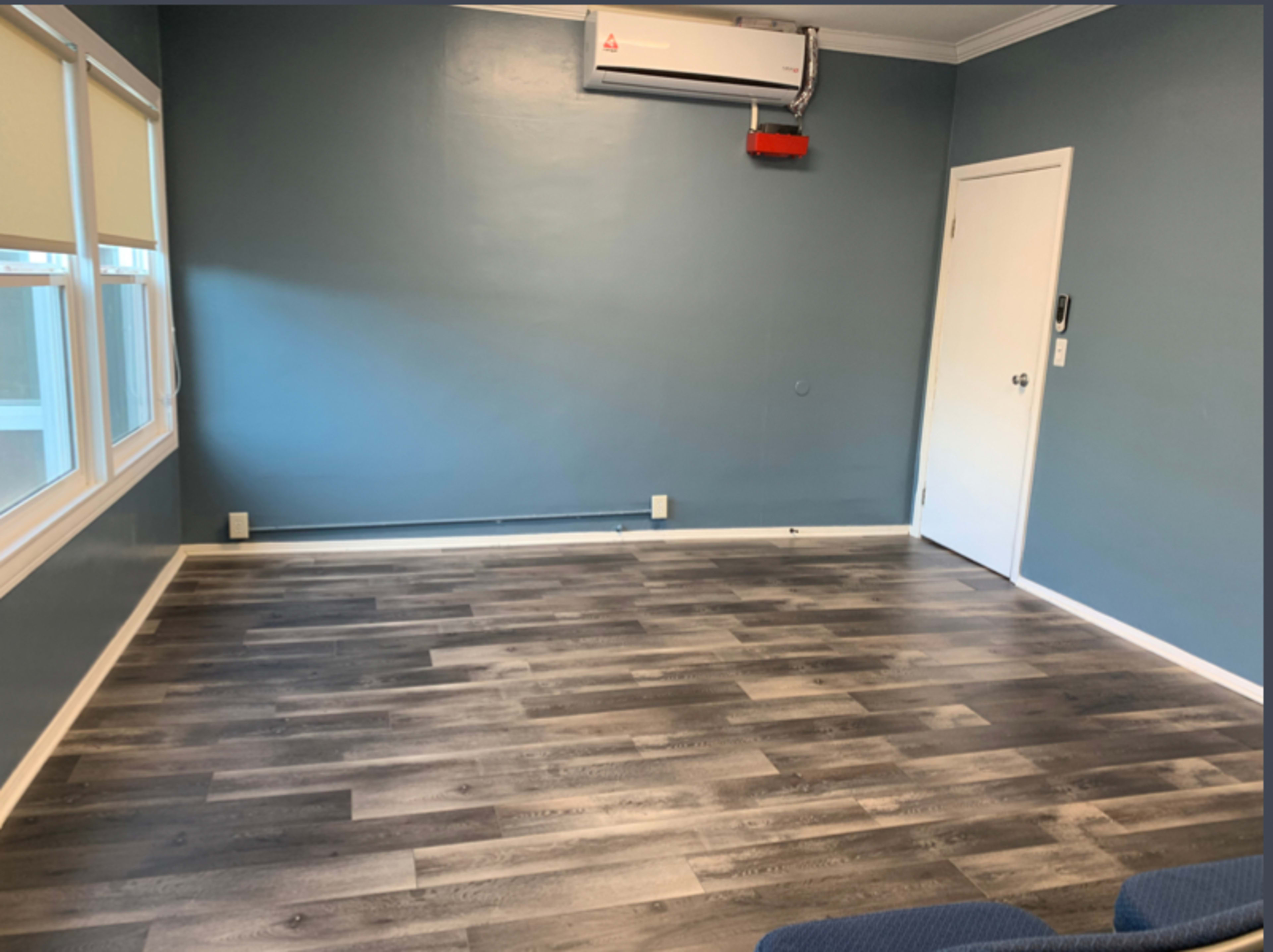 The image shows a sparsely furnished room with blue walls, wooden flooring, an air conditioning unit, and a single door.