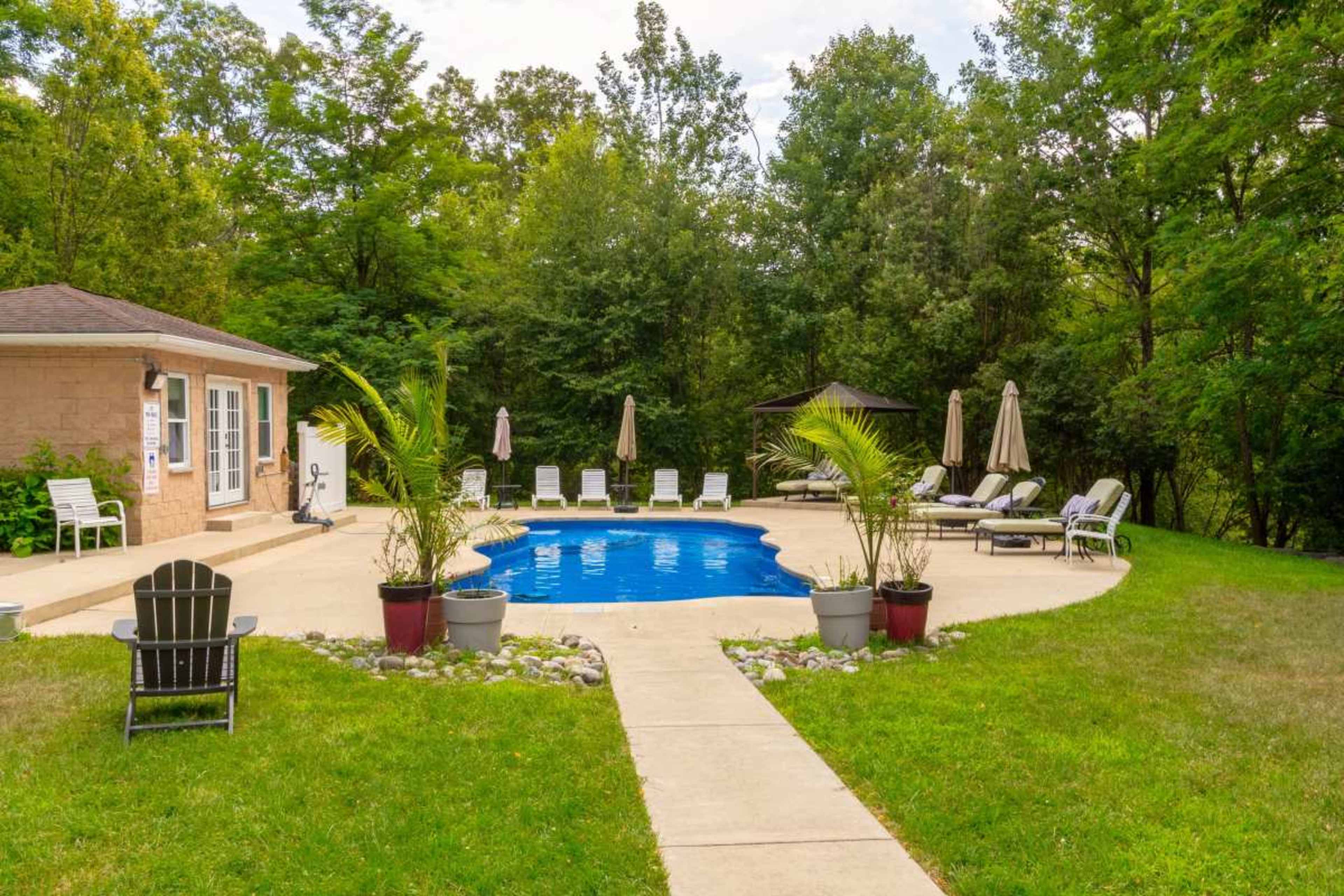 A blue swimming pool is surrounded by lounge chairs and umbrellas, with a patio area and greenery in the background.