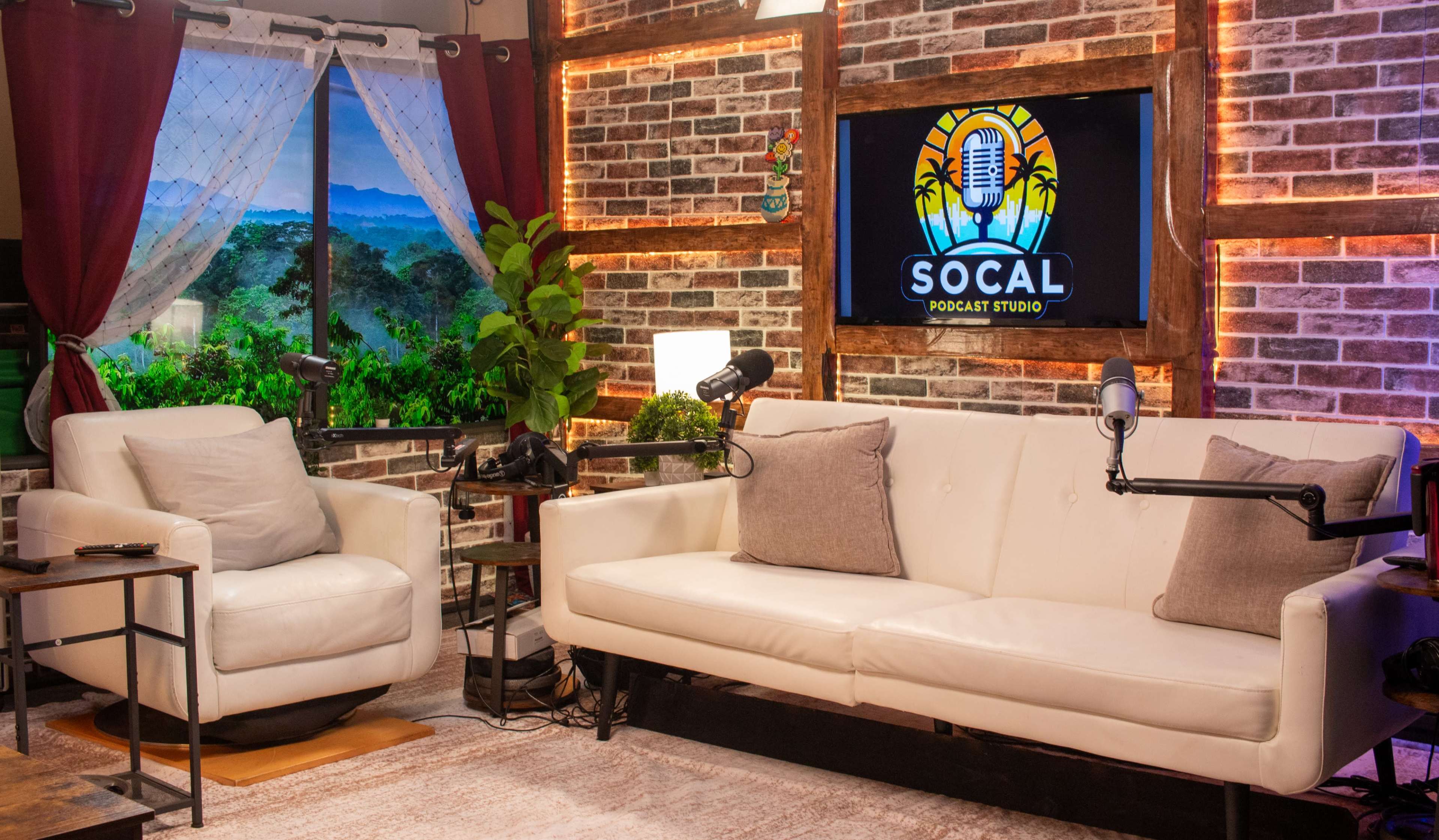 The image shows a podcast studio with a white sofa, two microphones positioned for recording, and a large screen displaying the "SO CAL PODCAST STUDIO" logo against a backdrop of brick walls and window curtains.