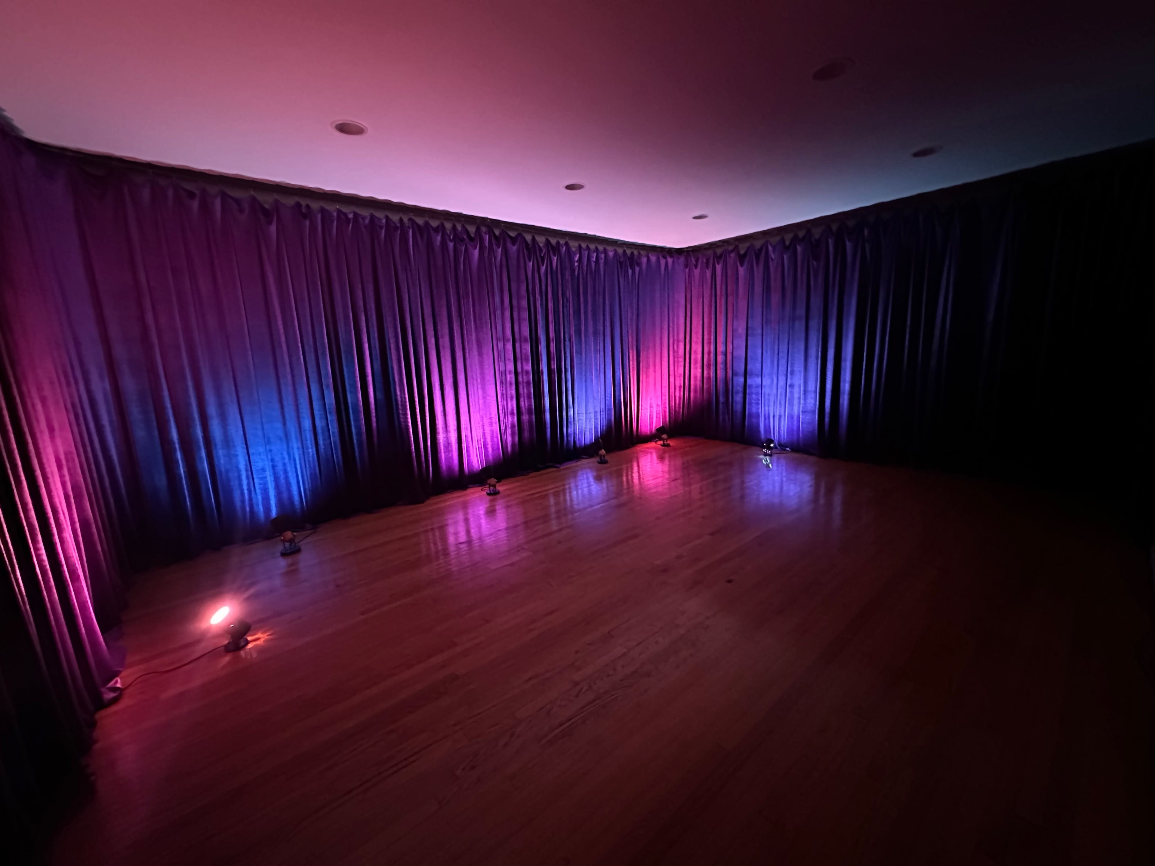 The image shows a dimly lit room with purple and pink curtains, illuminated by colored lights on the floor.