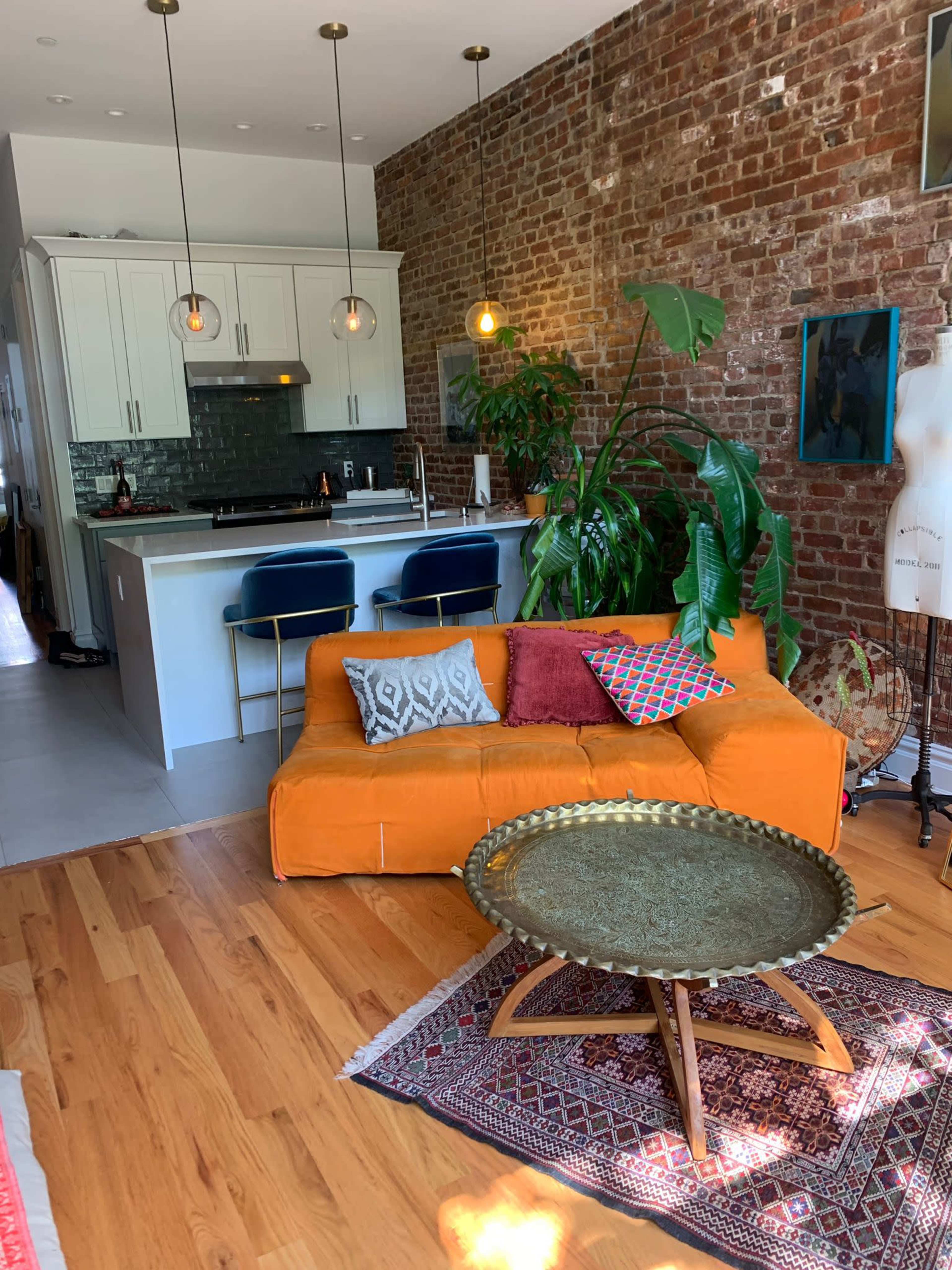 The image shows a cozy living space with an orange couch, a large round coffee table, a green plant, and a kitchen area with white cabinetry and modern fixtures.