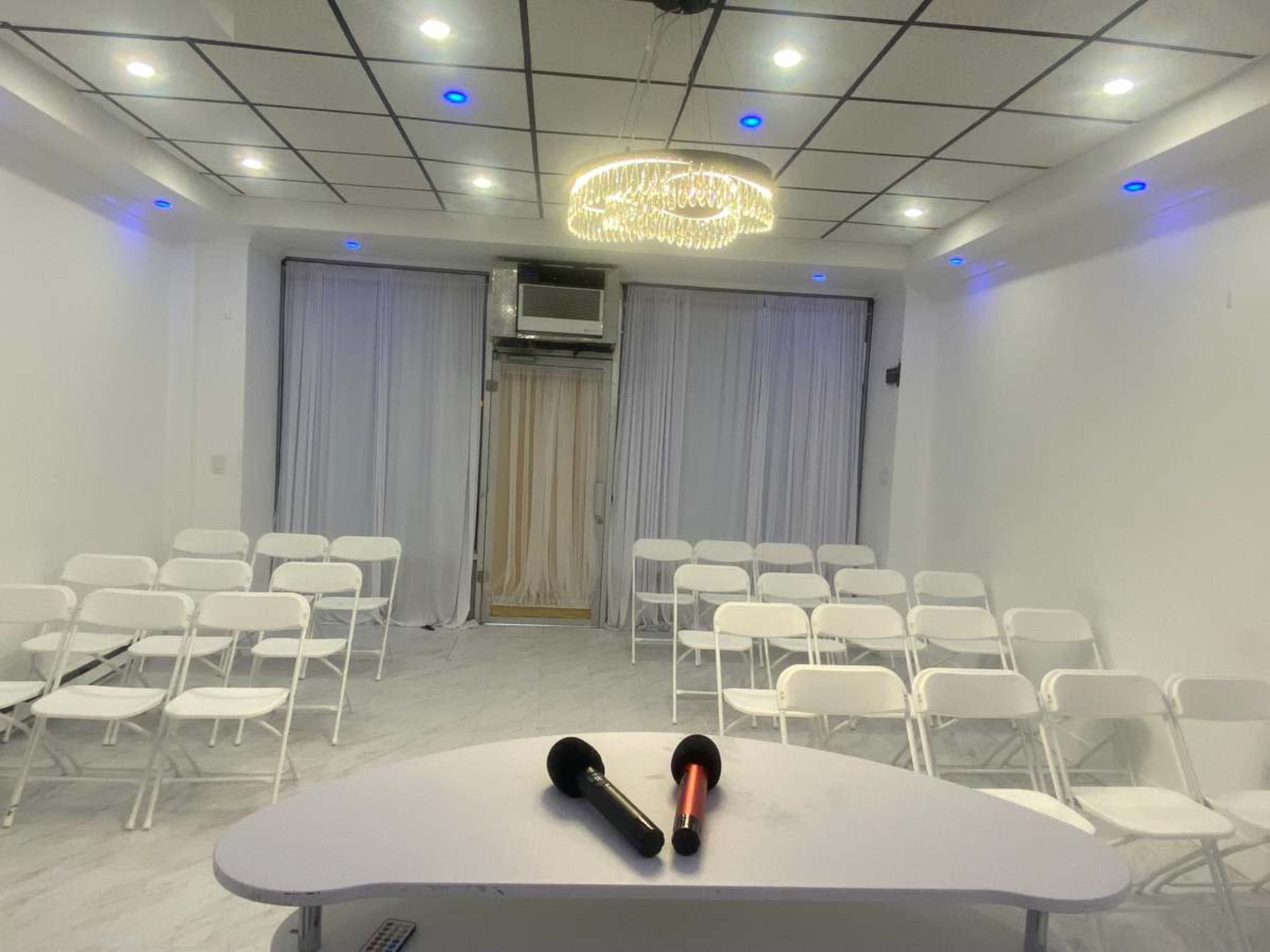 The image shows a small, well-lit event room with white chairs arranged in rows and a central table featuring two microphones.