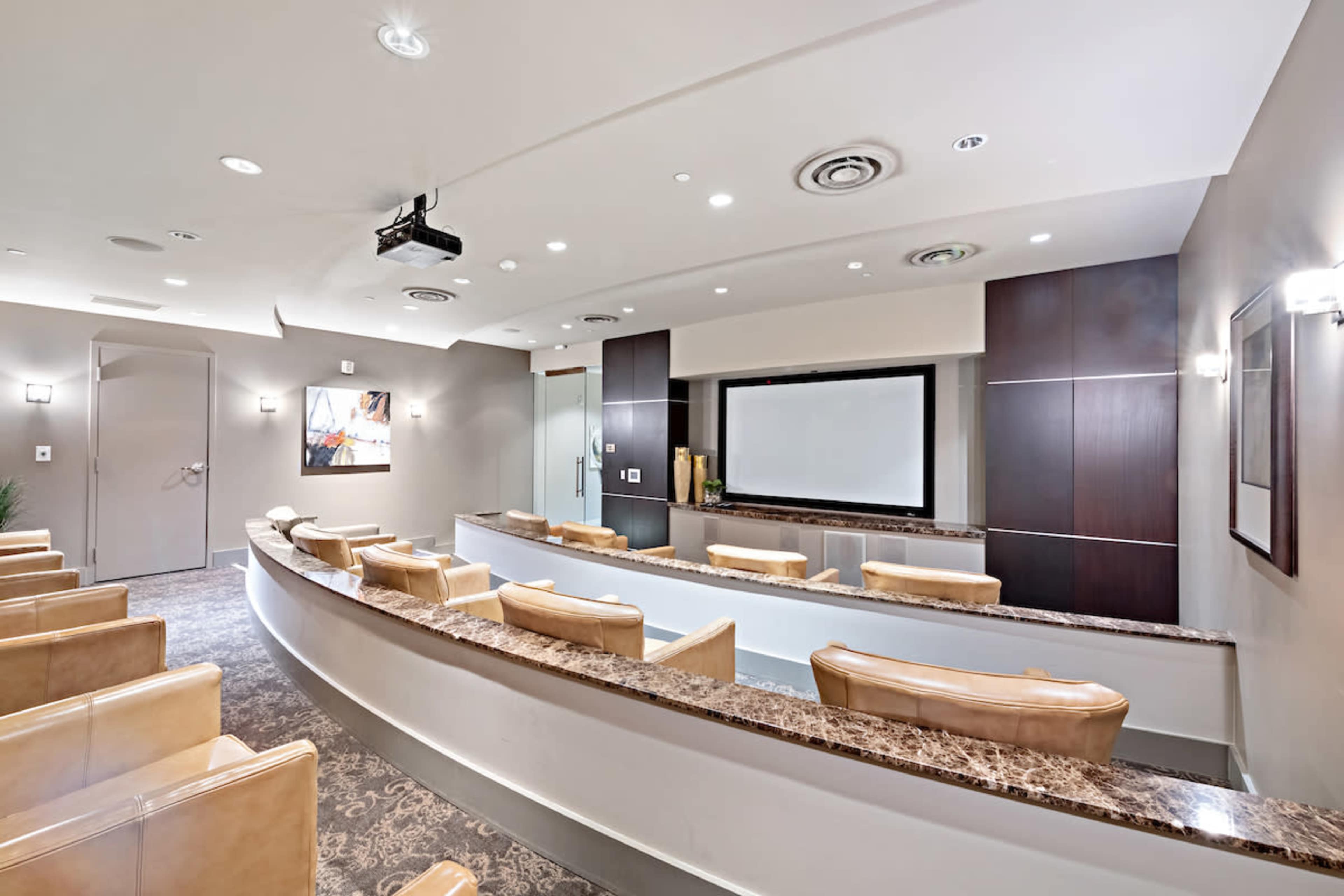 The image shows a modern conference room with tiered seating, a large screen at the front, and a projector installed in the ceiling.