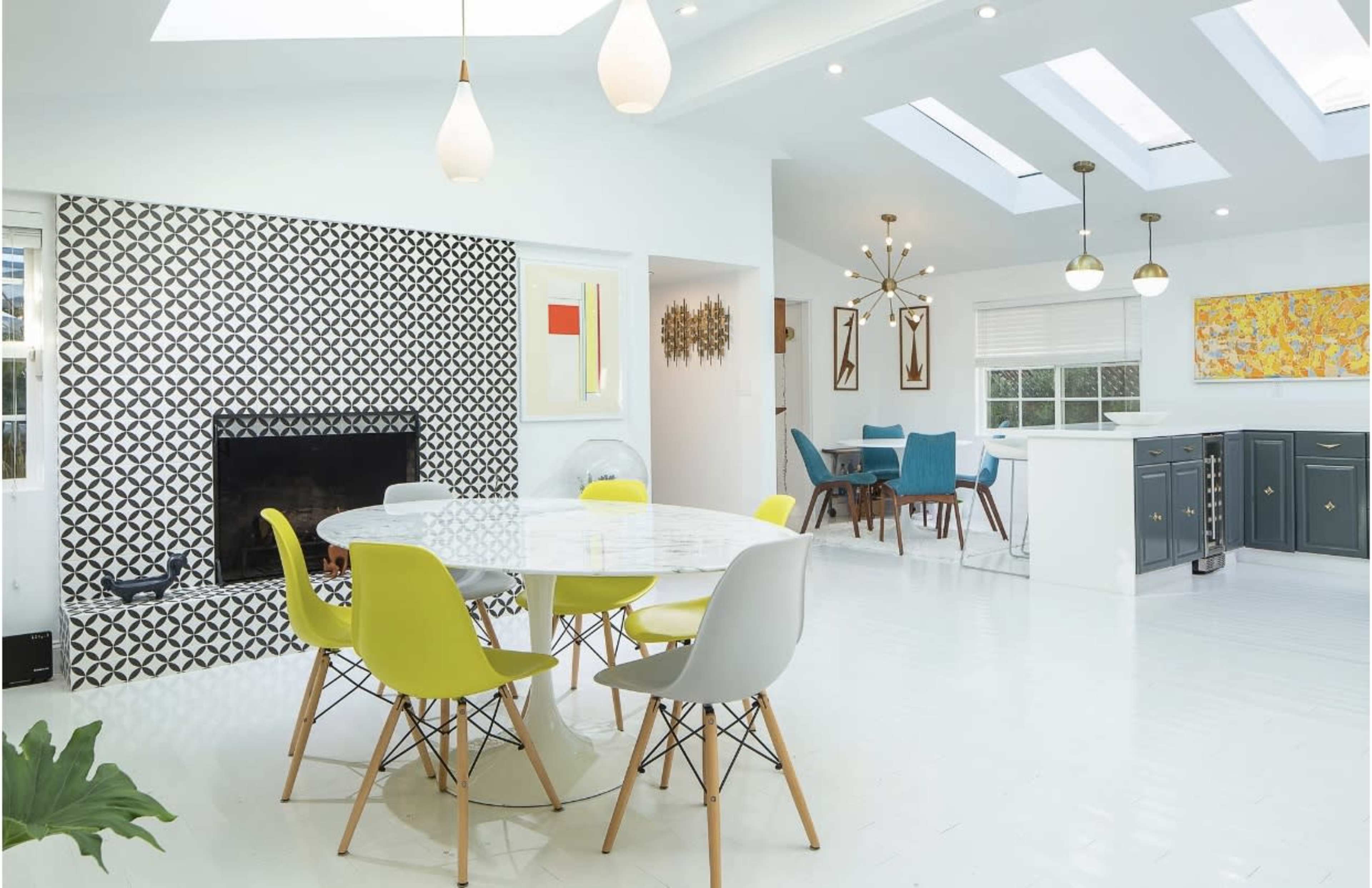 The image shows a modern dining area with a round marble table surrounded by colorful chairs, a patterned accent wall, and an open kitchen visible in the background.