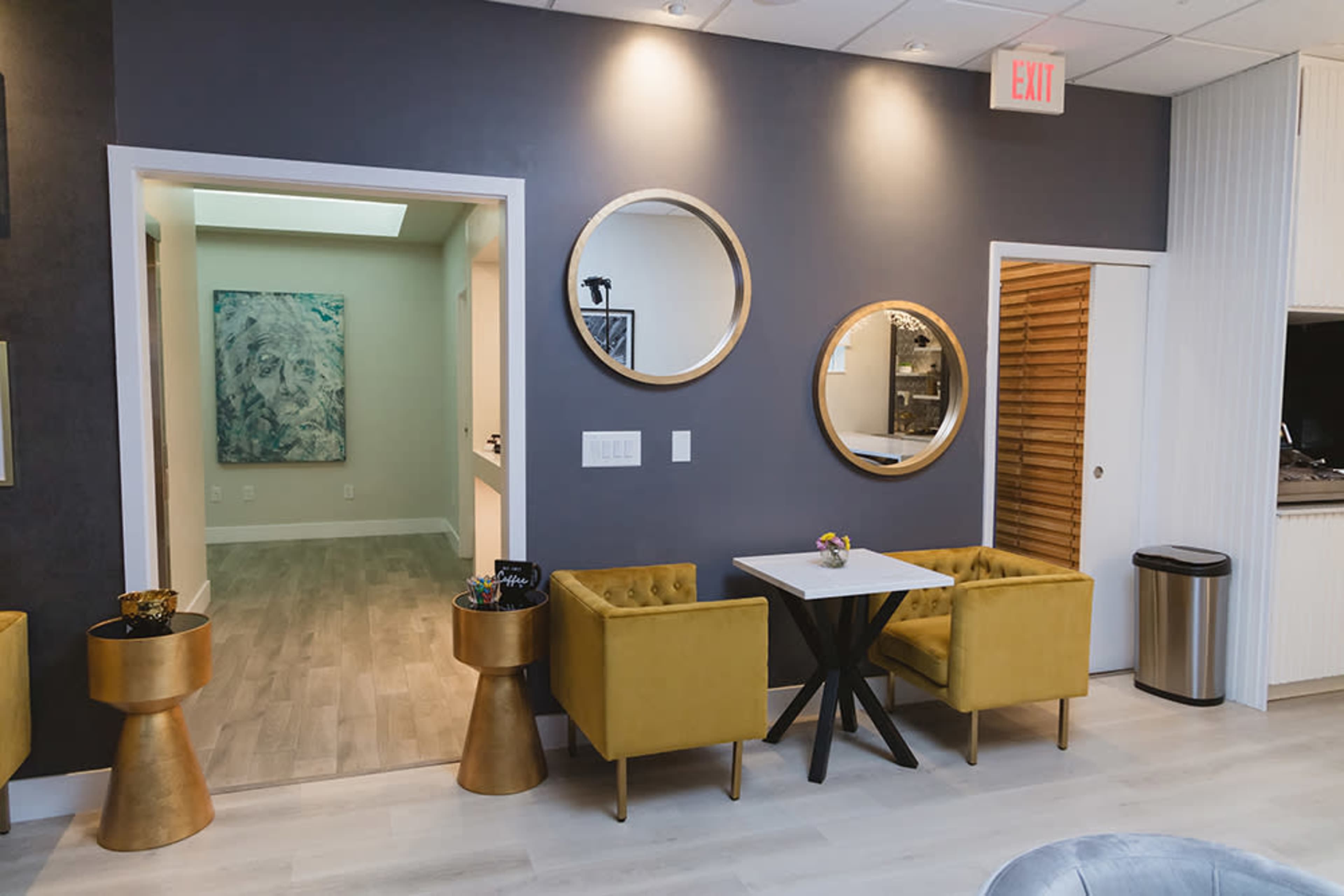 A modern interior featuring yellow chairs, a small black table, and gold accent tables against a gray wall, with mirrors on either side.