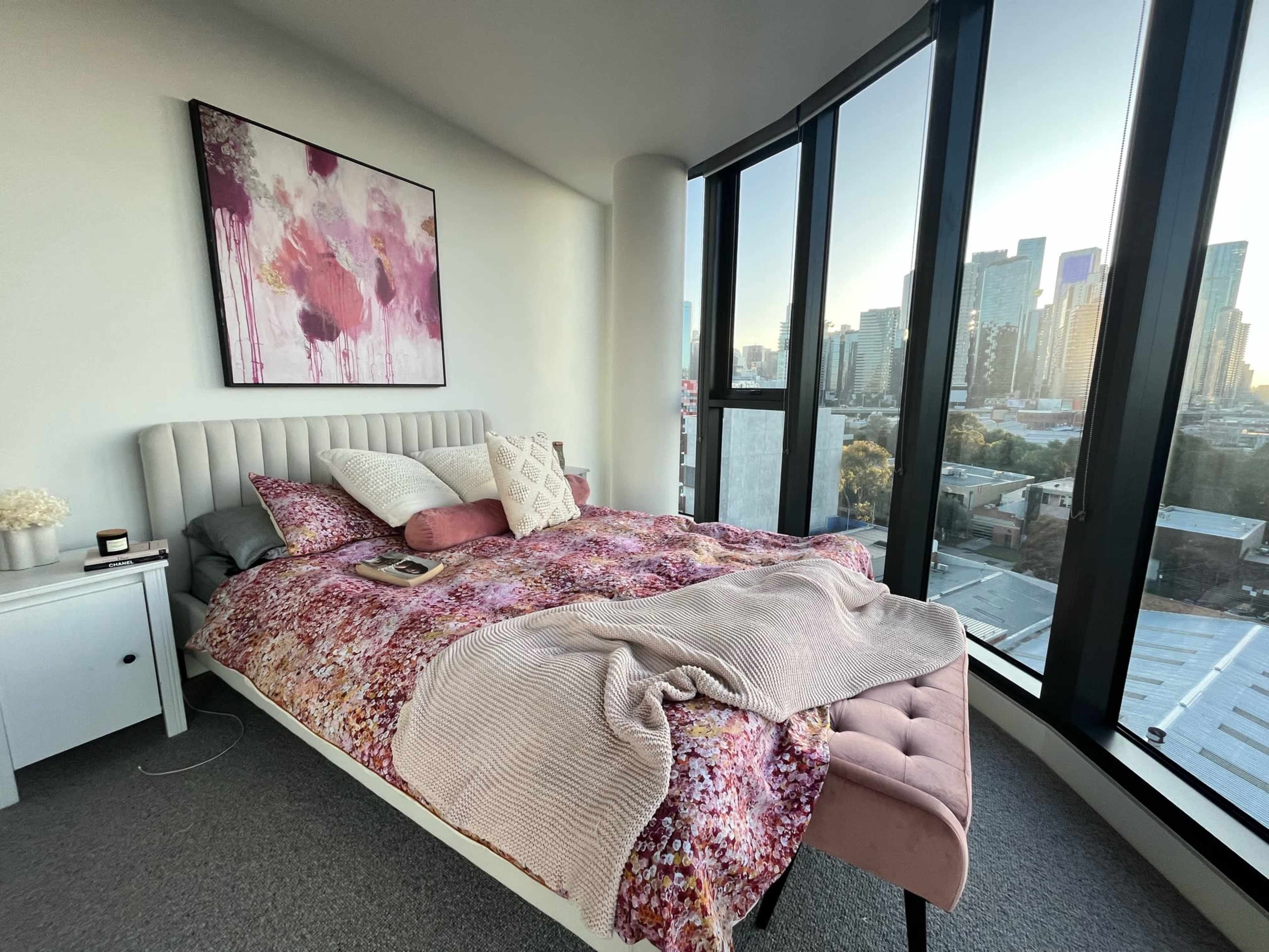 A bed with a patterned comforter and decorative pillows is positioned near large windows overlooking a city skyline.