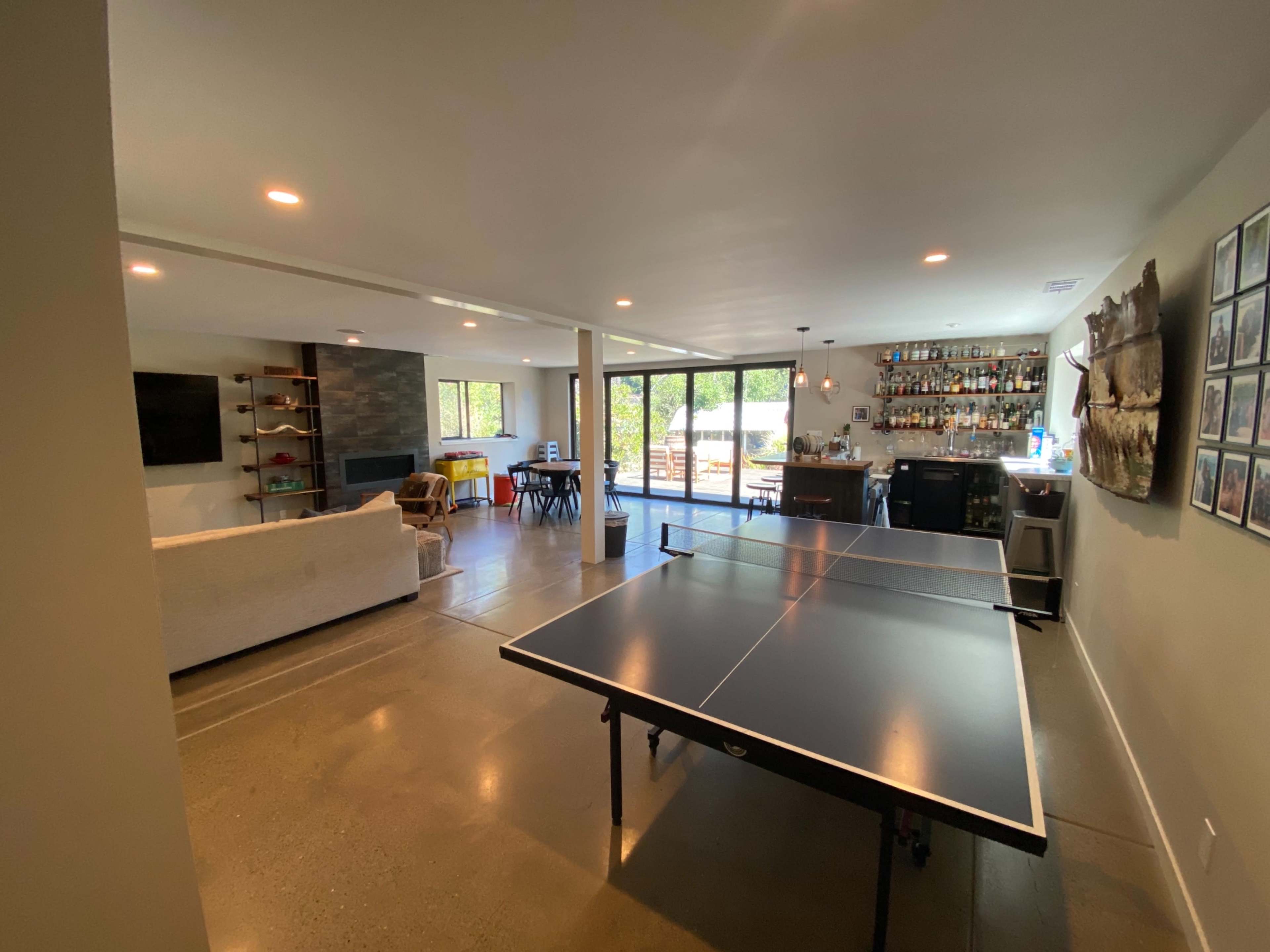 A spacious room featuring a ping pong table, a bar area with shelves of bottles, and large windows overlooking an outdoor space.