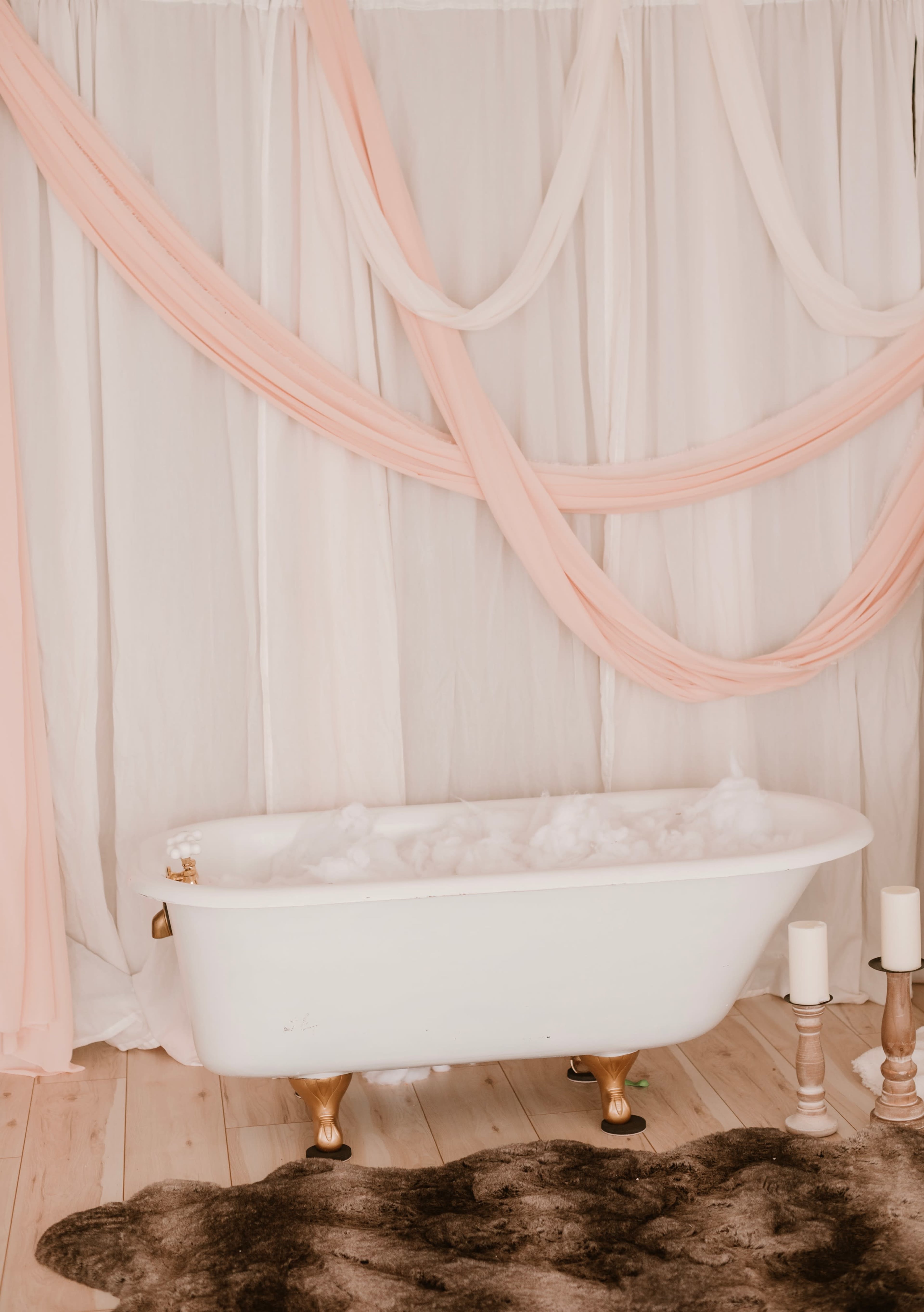 A vintage white bathtub filled with bubbles, placed against a backdrop of draped fabric and candles on a wooden floor.